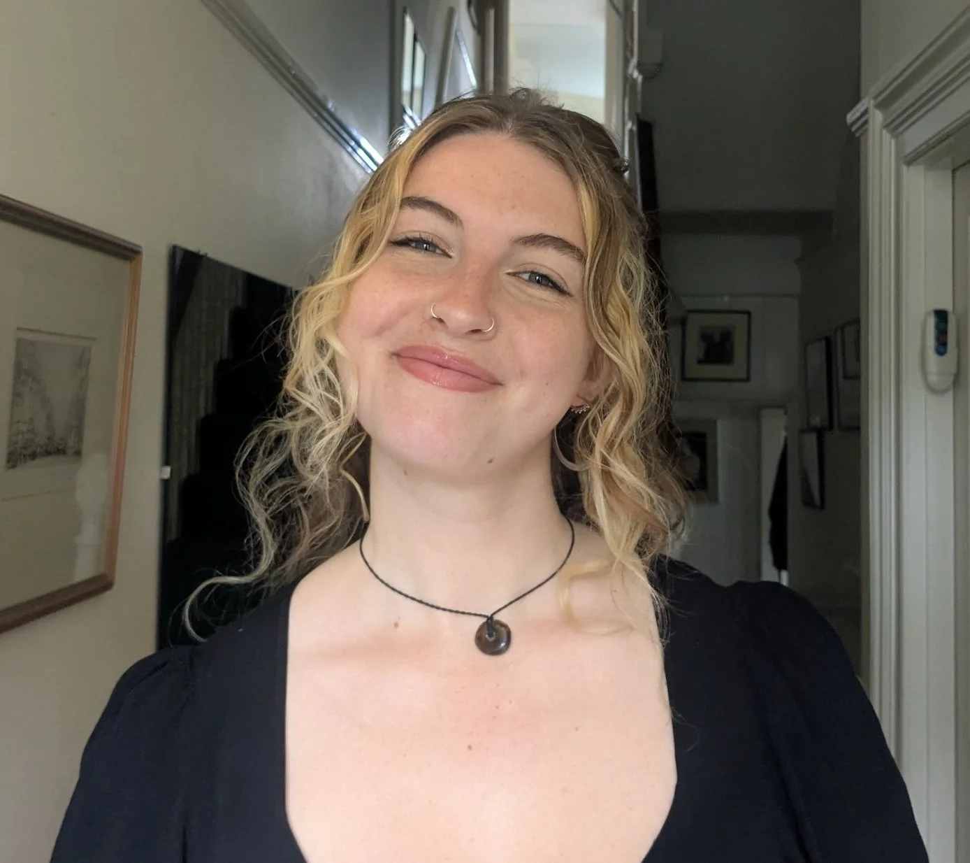 A smiling woman with curly blonde hair, wearing a black top and a black necklace with a round pendant, standing in a home interior.