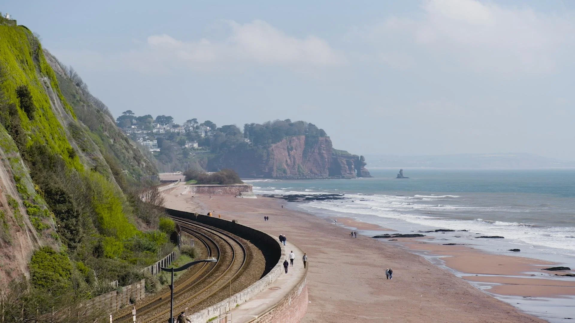 A scenic coastal view with a sandy beach, waves, green cliffs, and houses on top of the cliffs, with a curved railway track along the coast.