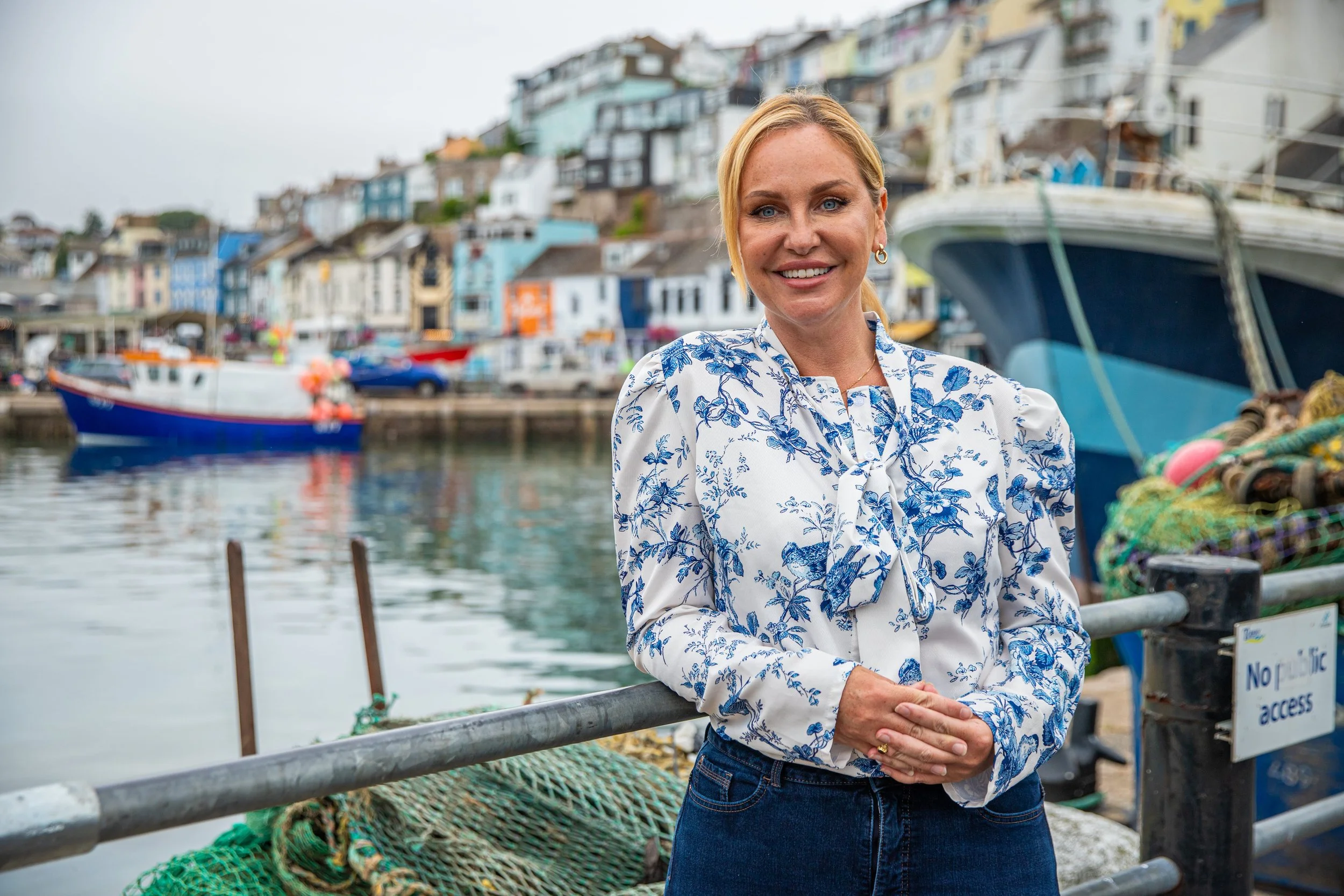 A woman with blonde hair wearing a white and blue floral blouse standing near a harbor with boats and colorful houses in the background.