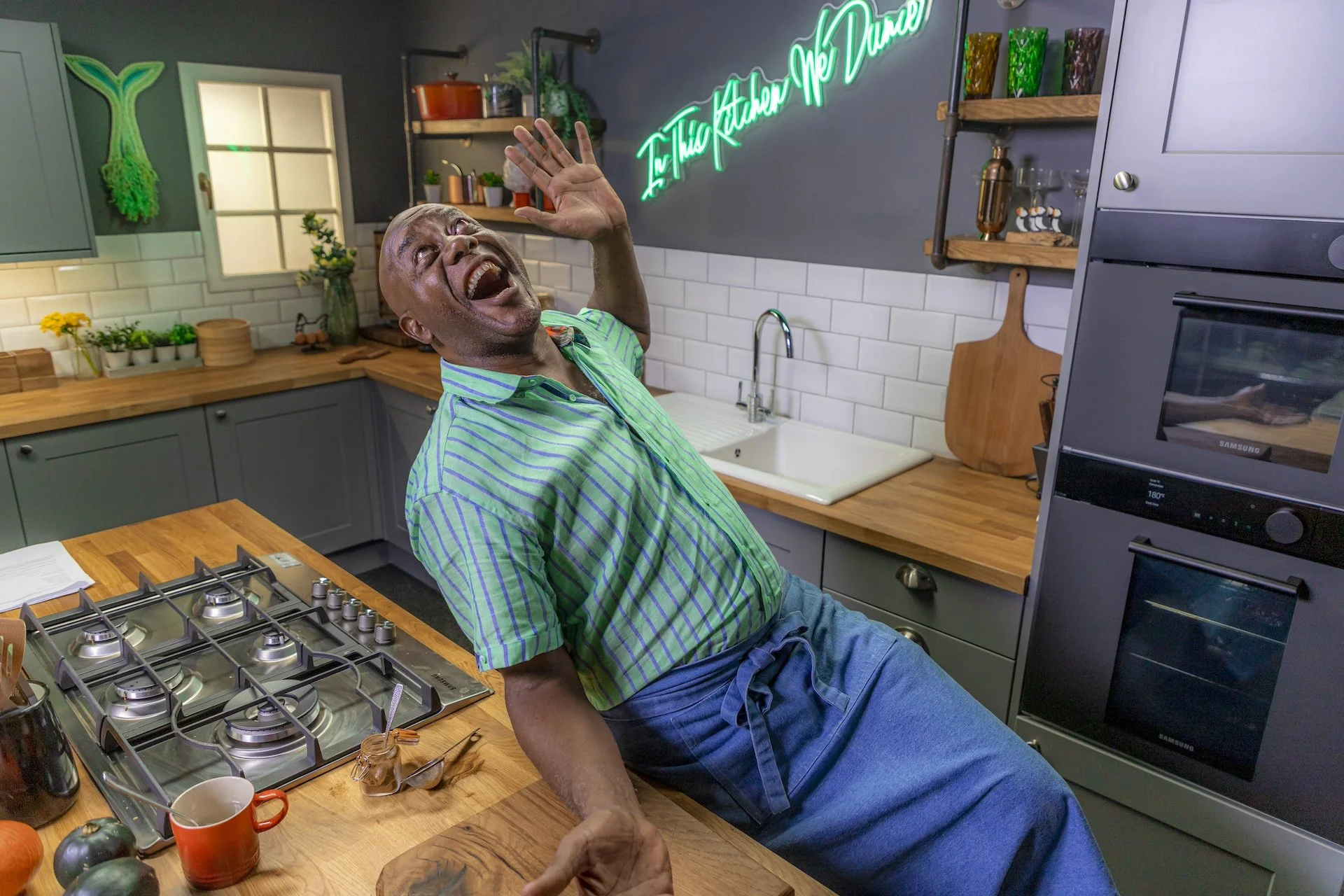A man with a big smile and an excited expression, wearing a green striped shirt, is leaning back in a kitchen with his hand raised in a joyous gesture. The kitchen has gray cabinets, wooden countertops, a gas stove, and decorative items including a neon sign that reads 'The Kitchen for Me Dine'. There are plants, glasses, and colorful cups on open shelves. The scene appears lively and cheerful.