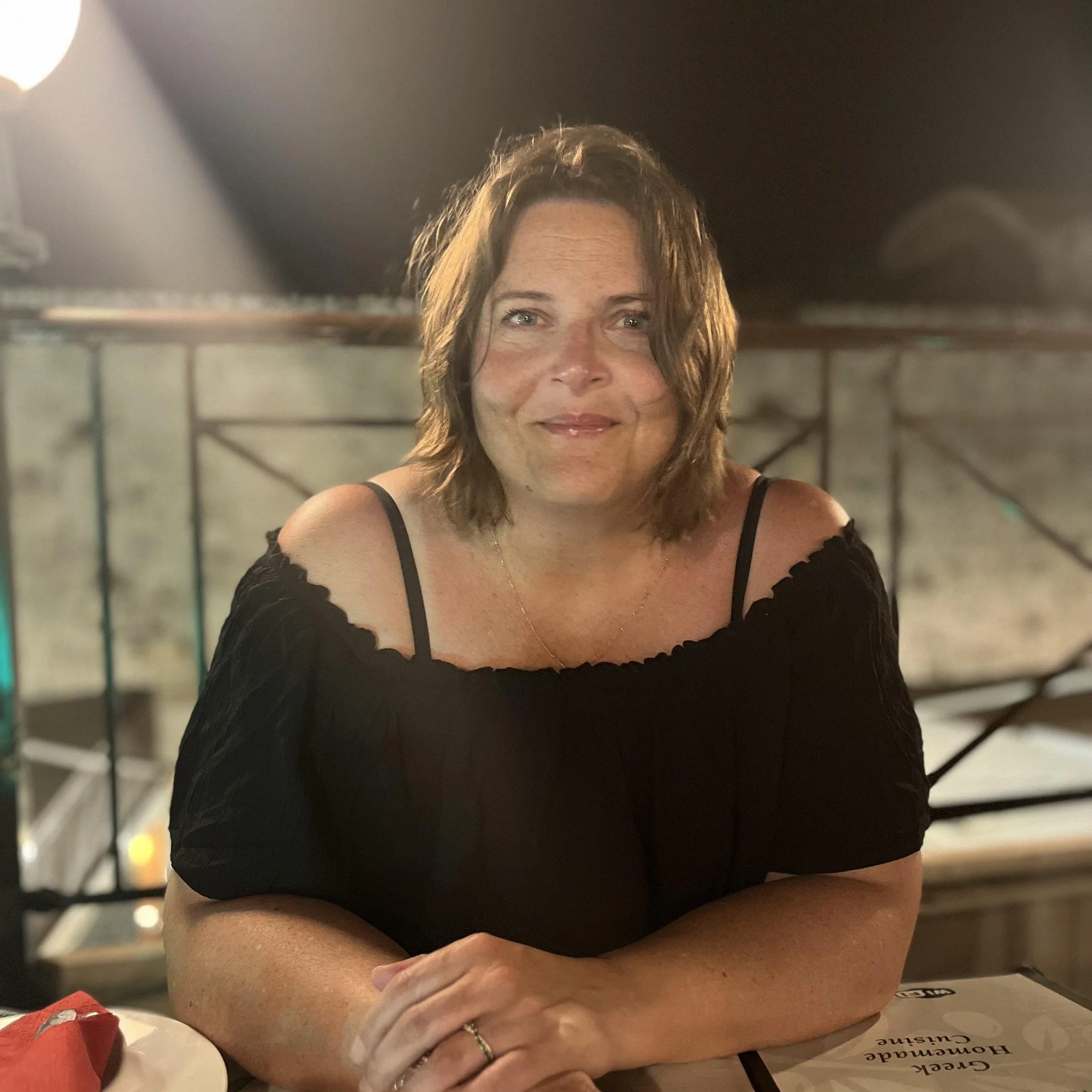 A woman with wavy brown hair and fair skin seated at a table, smiling, with her elbows resting on the surface, wearing a black off-shoulder top.