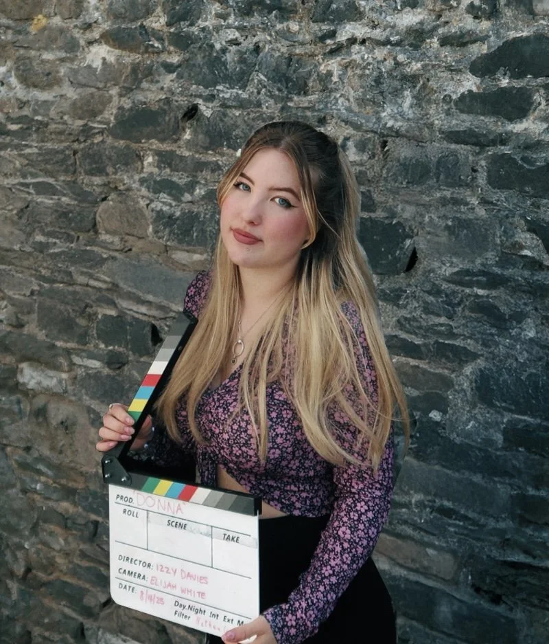 Young woman with long blonde hair and smiles holding a film slate, standing in front of a stone wall.