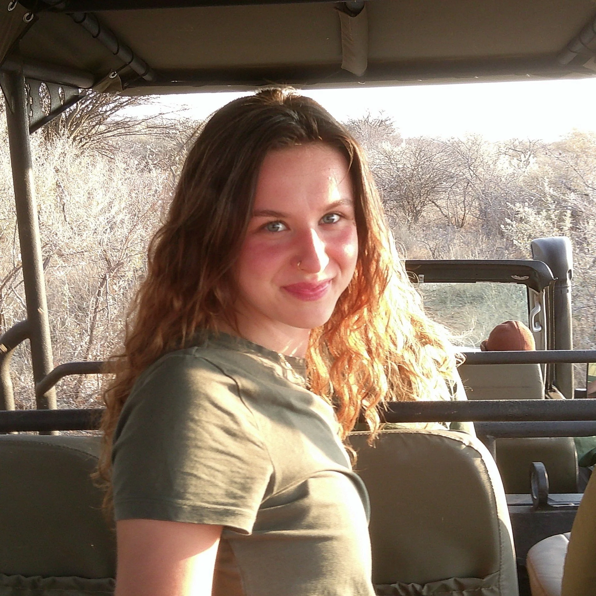 A woman with long, curly red hair on a golf cart during daylight, with trees in the background.