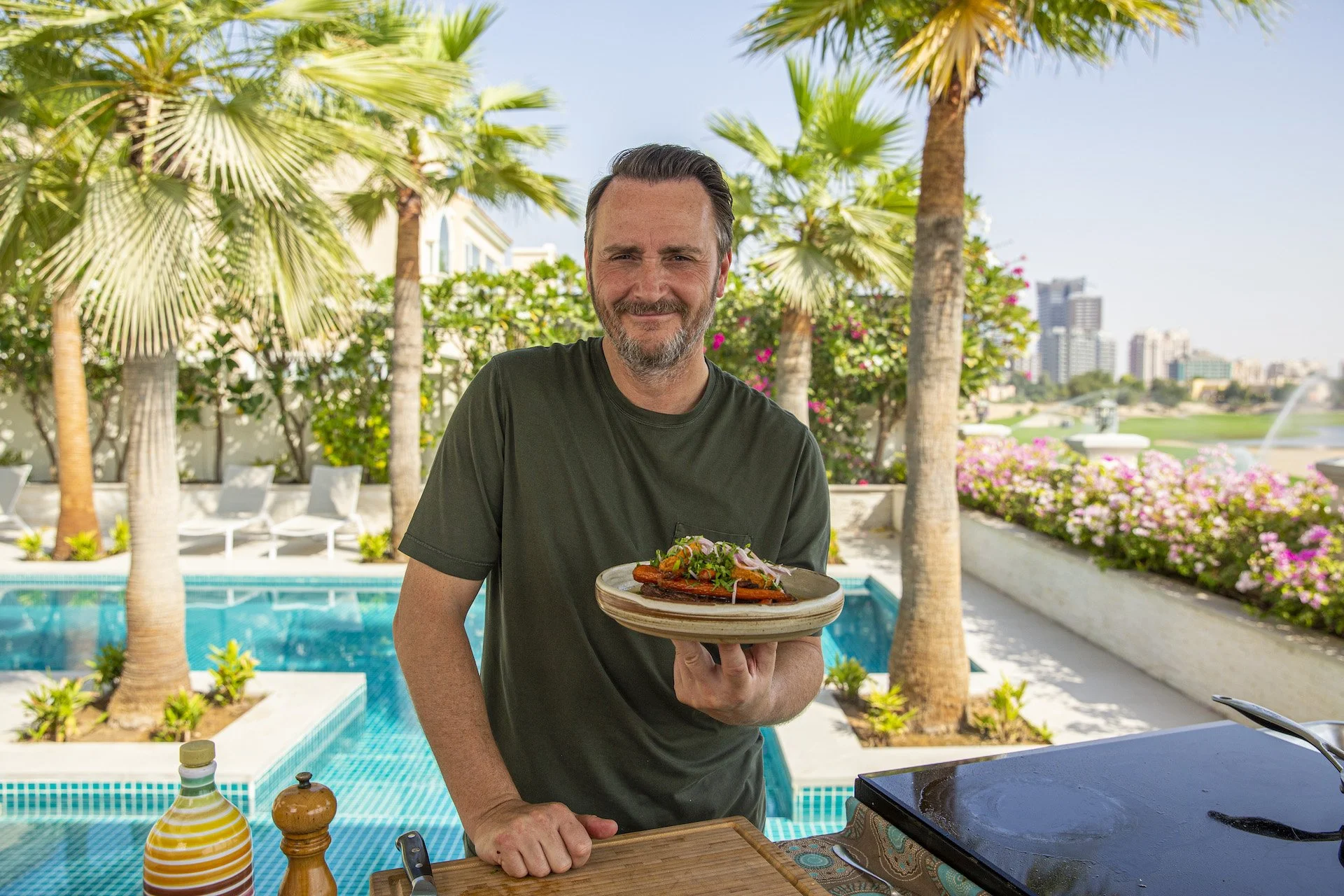A man with dark hair and a beard is standing outdoors near a swimming pool, holding a plate of food. The background features palm trees, pink flowers, and a city skyline with tall buildings.