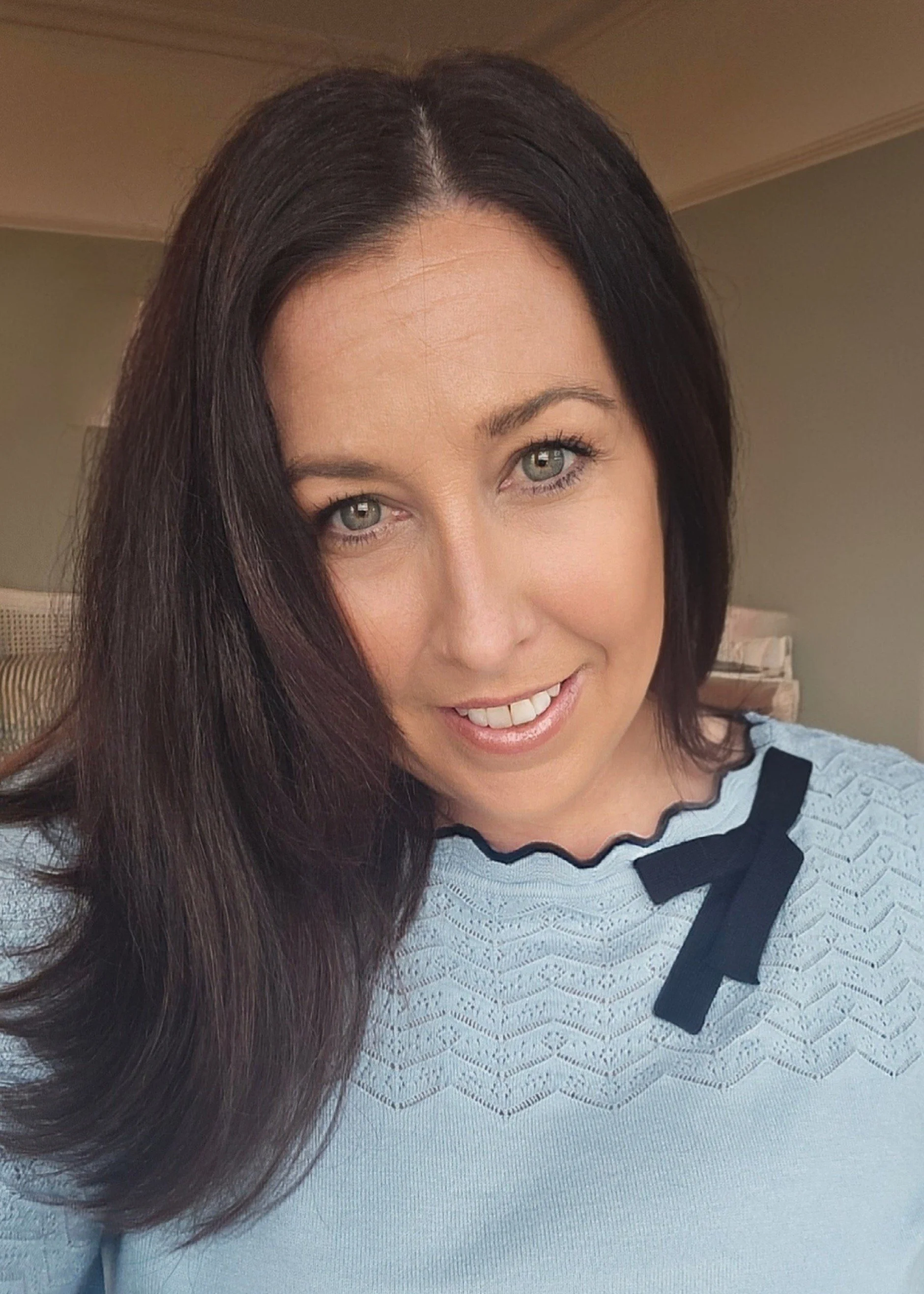 A woman with long dark hair and green eyes smiling while taking a selfie indoors, wearing a light blue top with black lace and bow details.