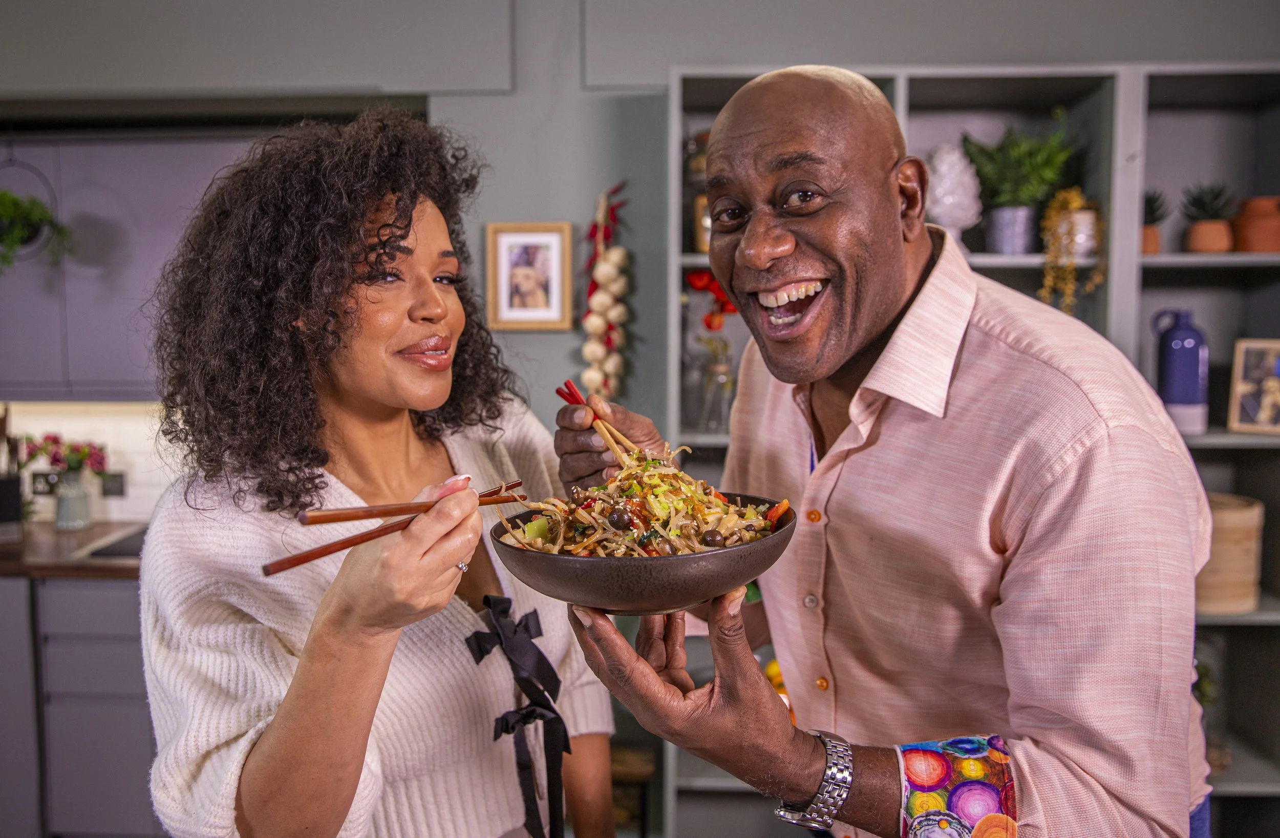A woman and a man share a bowl of stir-fried noodles topped with vegetables. The woman is wearing a white sweater and the man is wearing a peach shirt and colorful shorts. They are smiling in a modern kitchen with shelves and potted plants in the background.