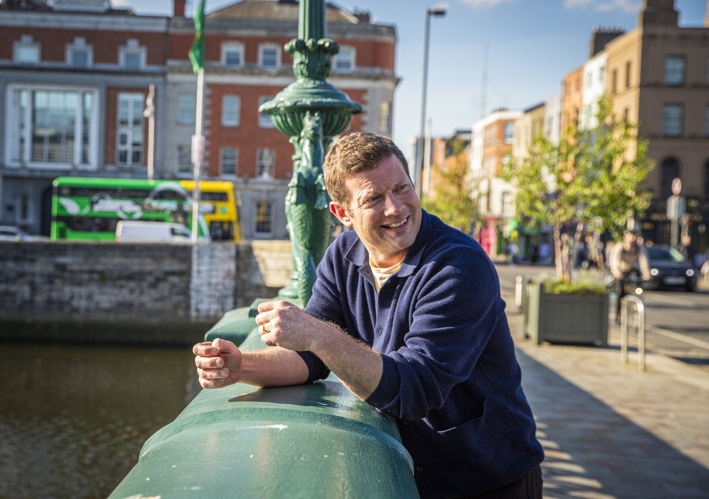 A man leaning on a green bridge railing, smiling, with buildings, trees, and a bus in the background on a sunny day.