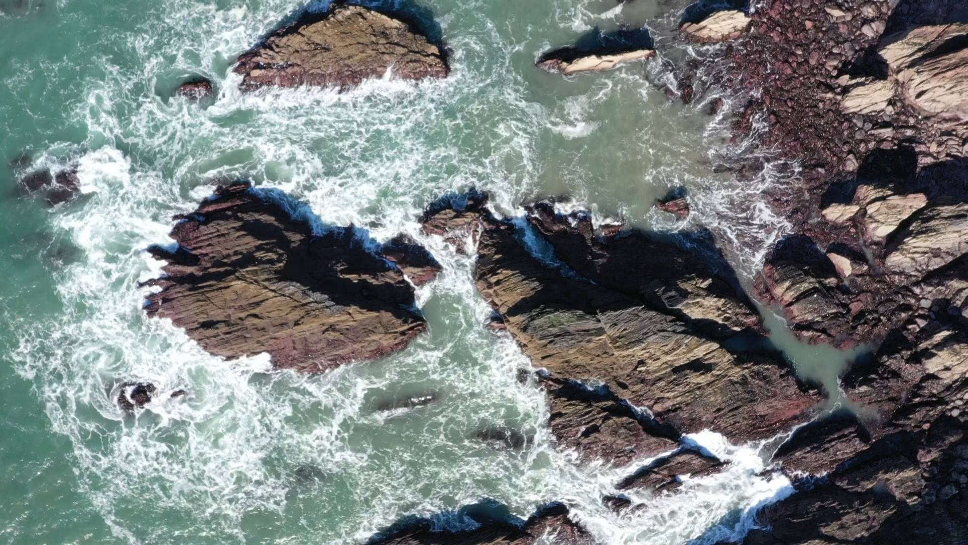 An aerial view of ocean waves crashing against large dark brown rocks along a shoreline.