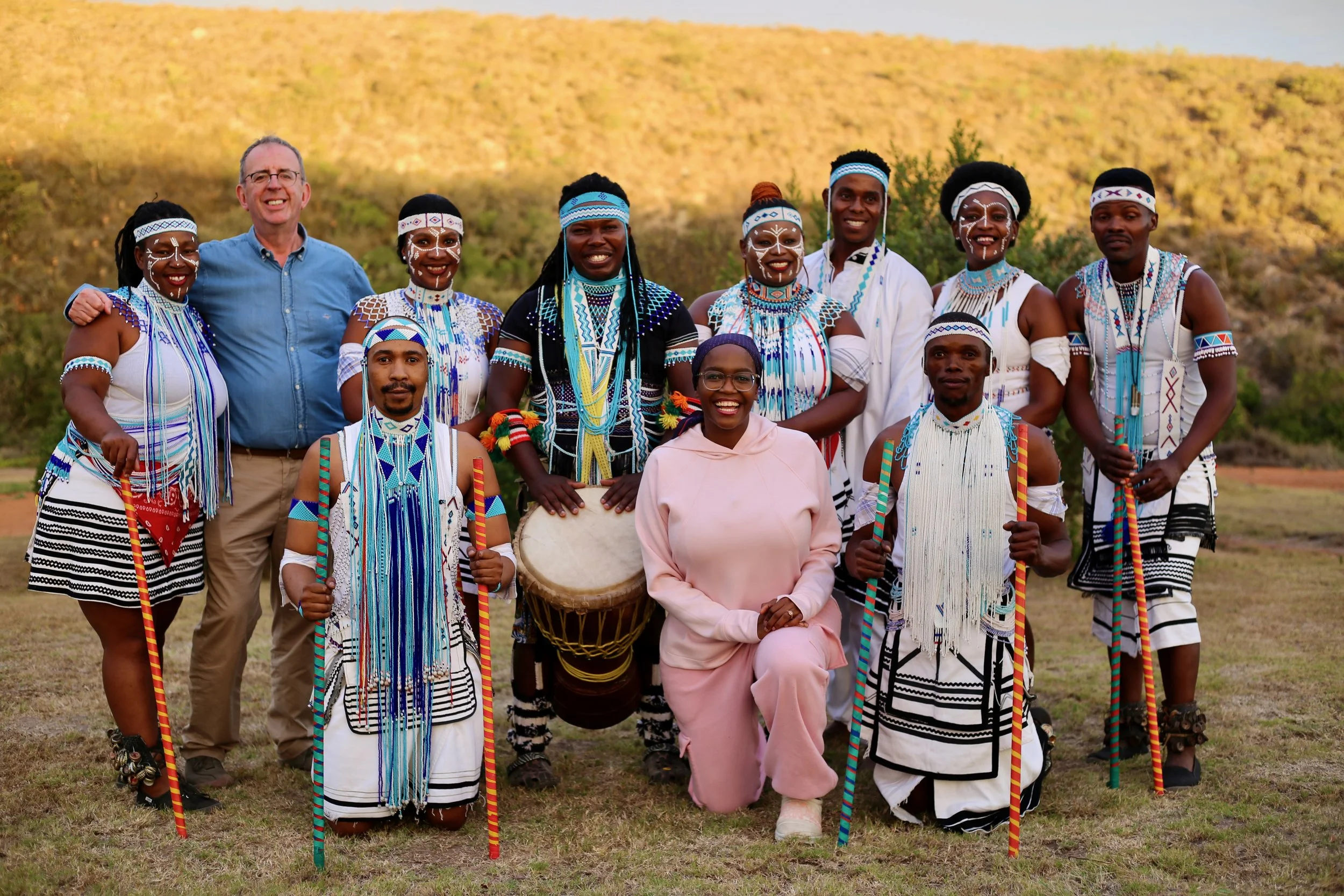 Group of people, including men and women, dressed in traditional Maasai attire with colorful beaded jewelry and clothing, standing outdoors on grass with a hilly landscape in the background. One person is holding a drum, and a man in a blue shirt stands among them.