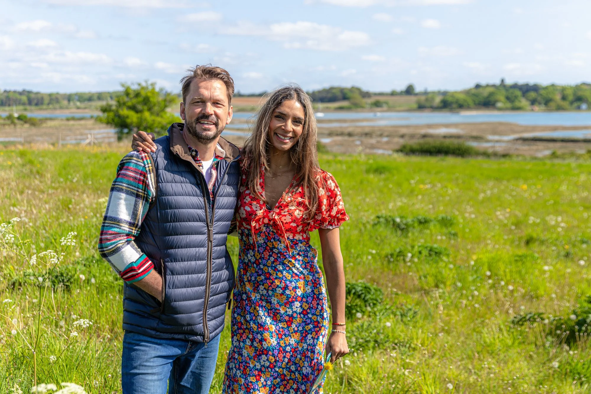 A man and a woman standing together outdoors, smiling, with a landscape of water, trees, and blue sky in the background.
