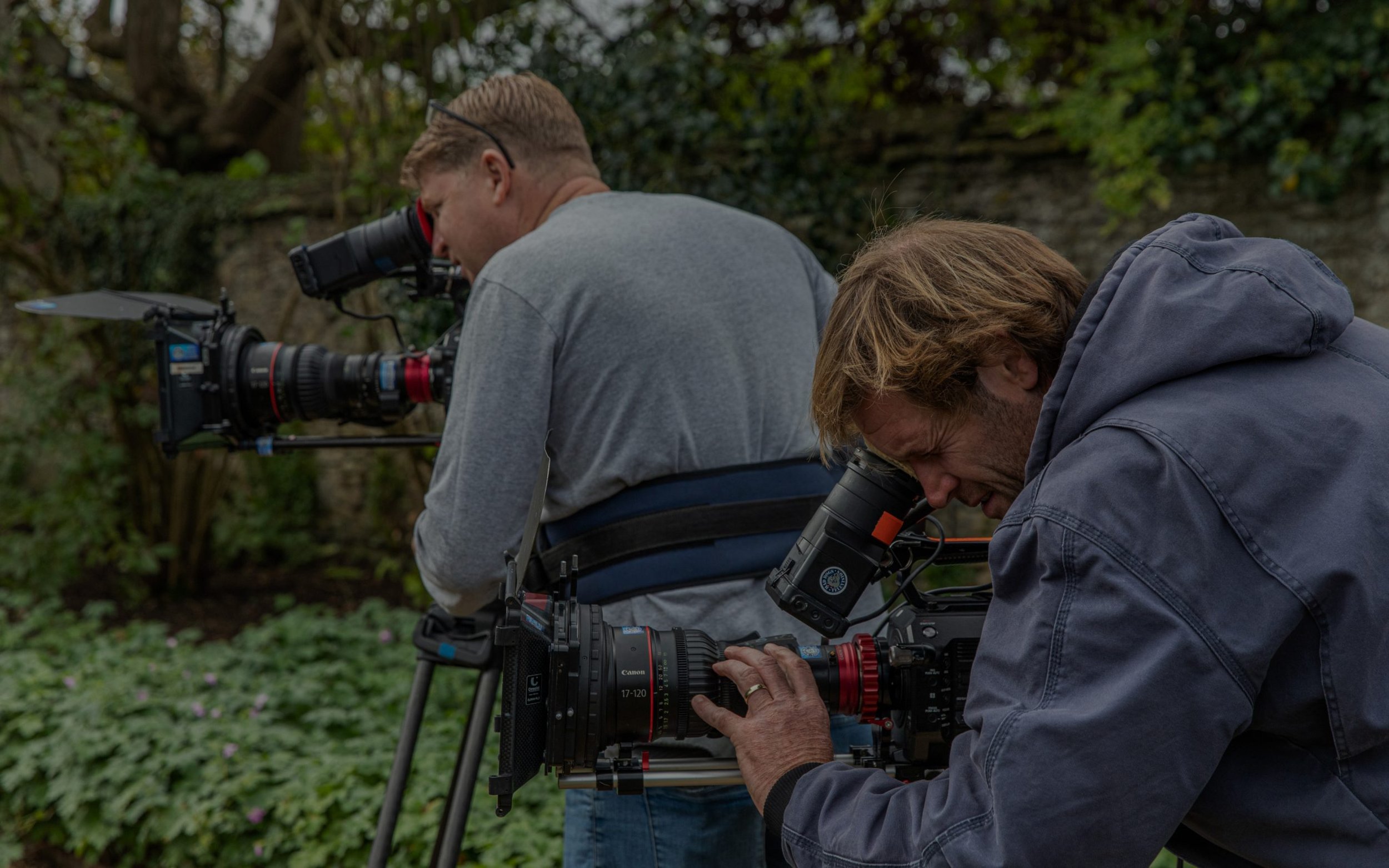 Two men are working with professional camera equipment outdoors in a natural setting with green foliage in the background.