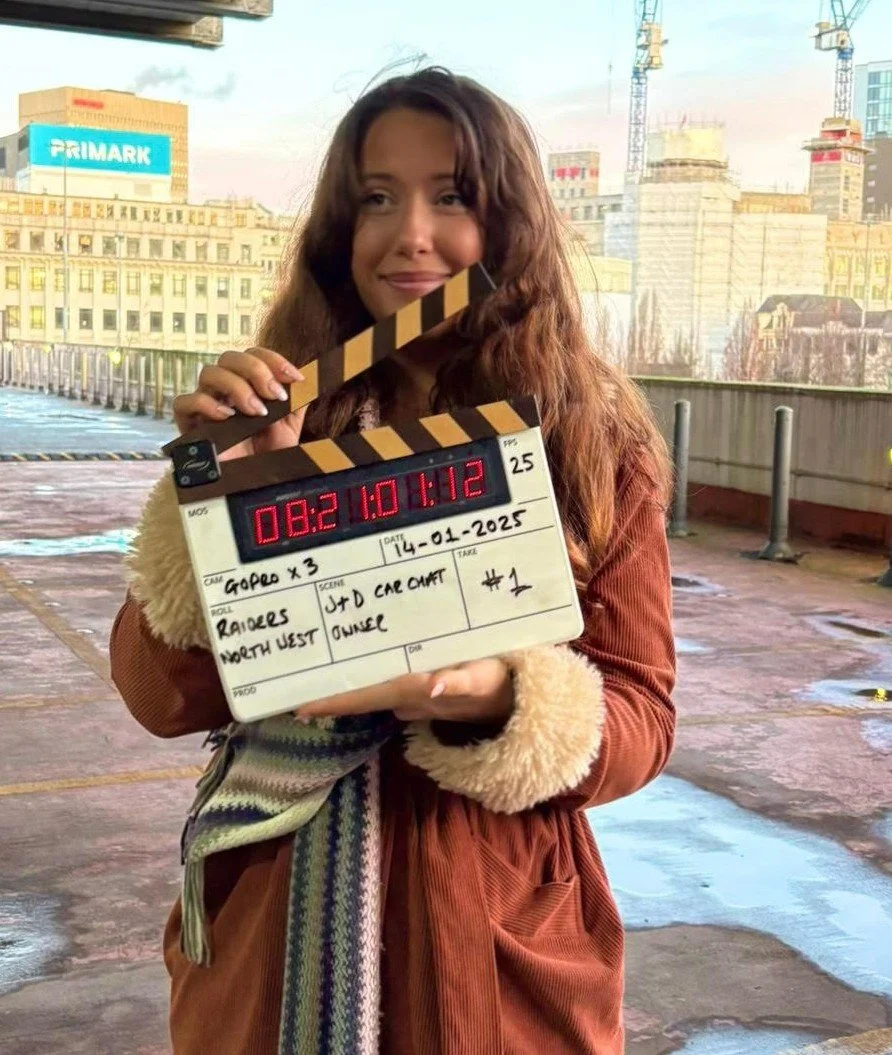 Young woman holding a film clapperboard outdoors with city buildings and cranes in the background.