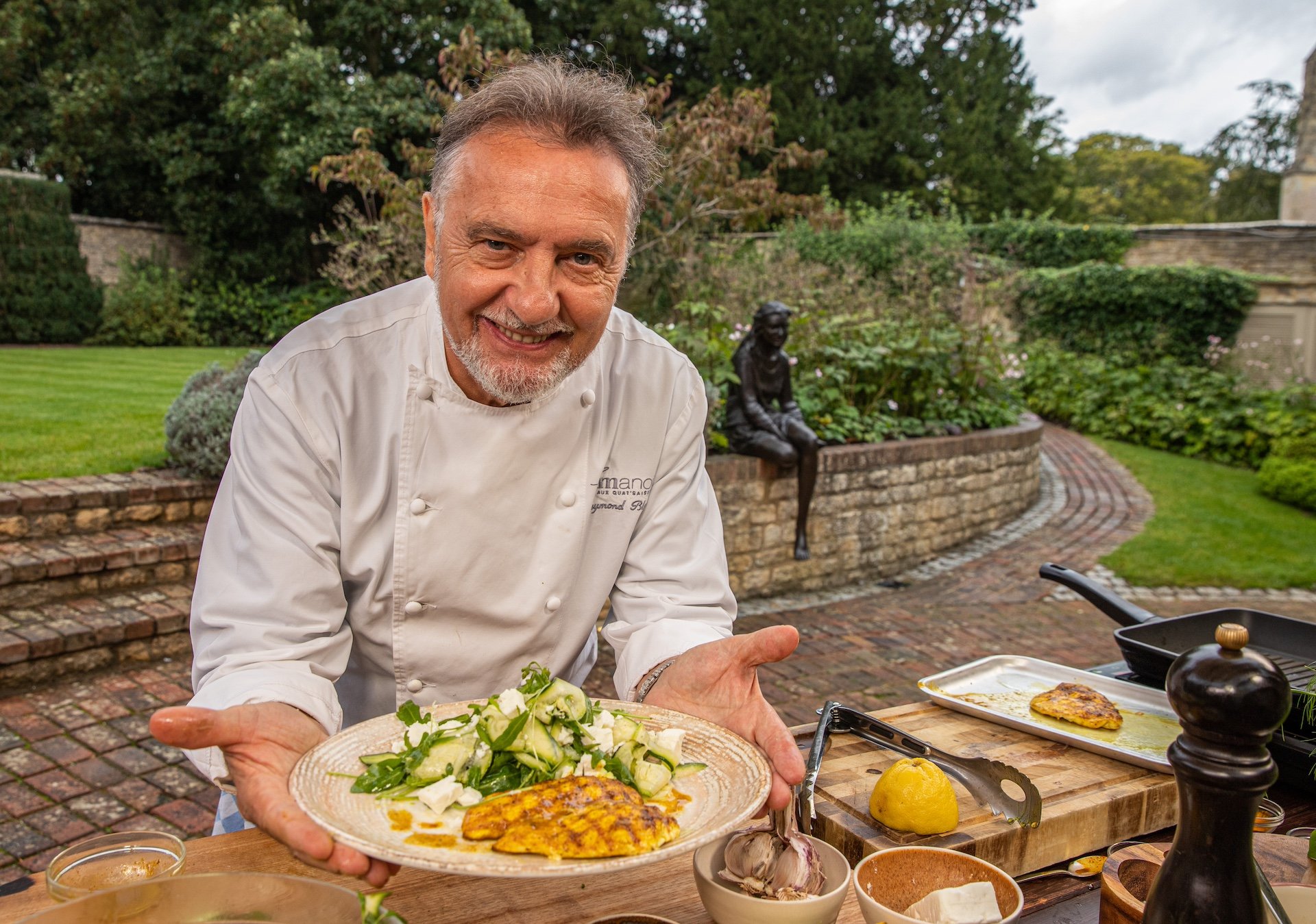 A male chef holding a plate of salad and grilled fish outdoors in a garden patio with greenery and brick pathway.