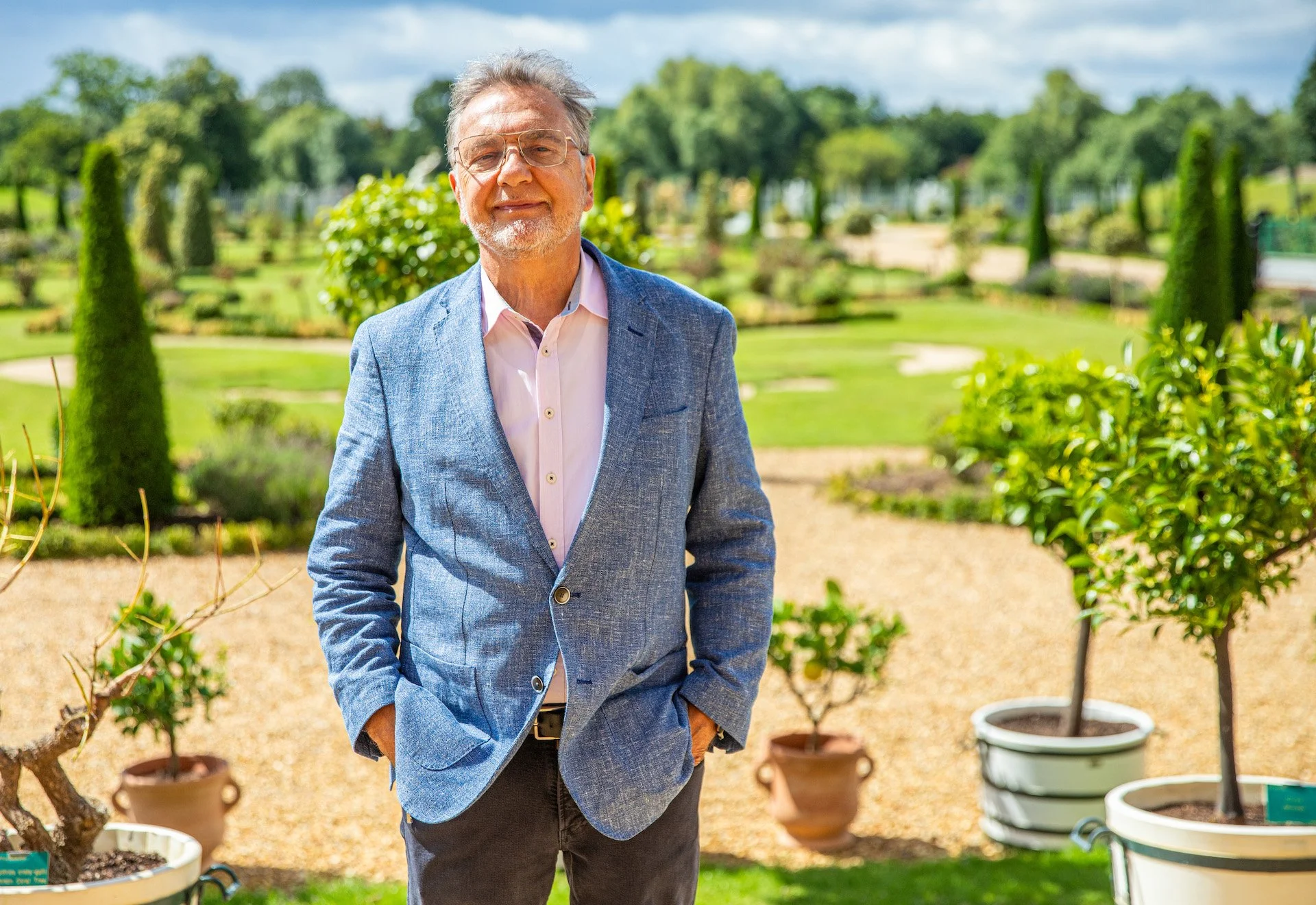 A man in a light pink shirt and blue blazer standing outdoors in a garden with trees and potted plants in the background, smiling at the camera.