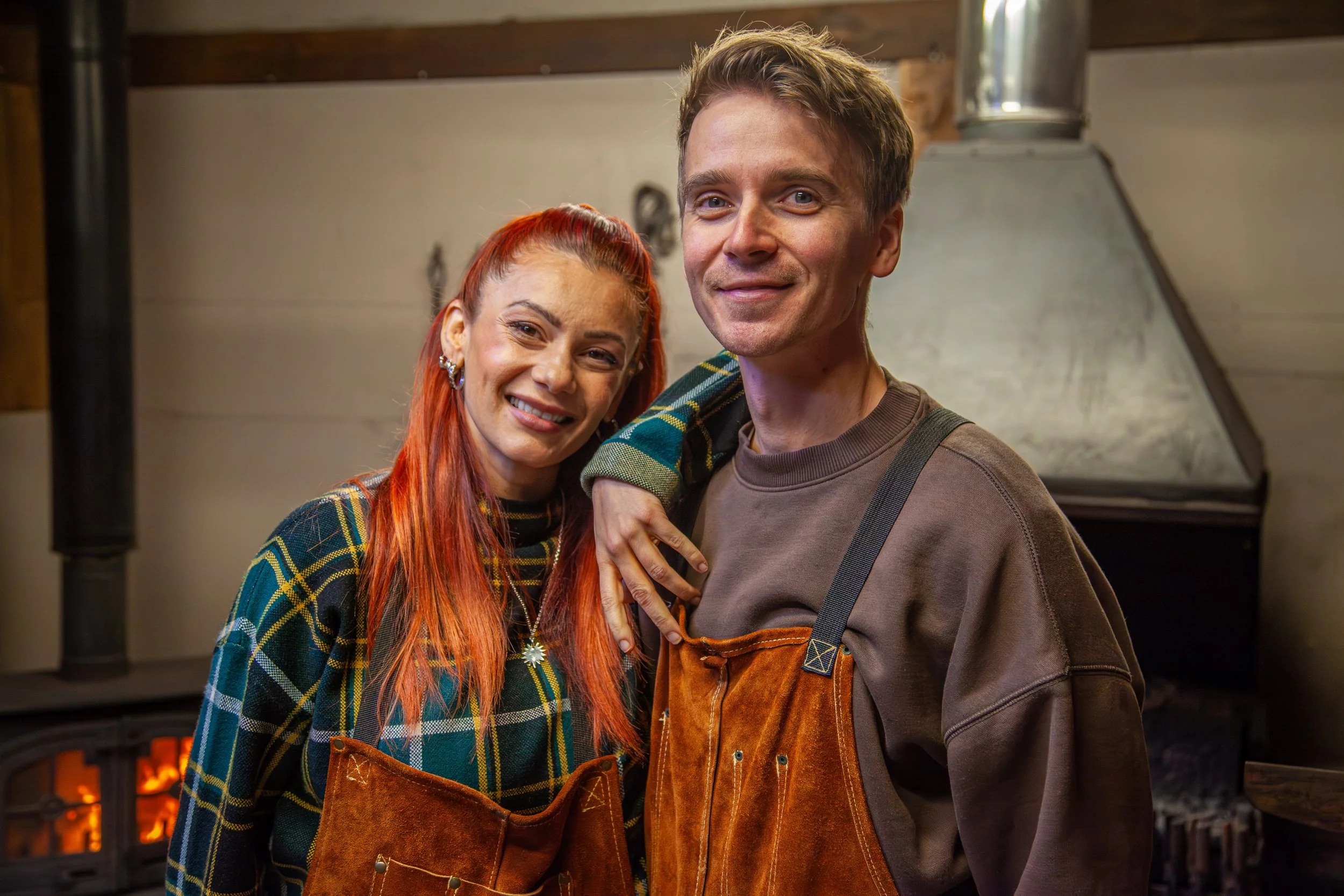 A smiling woman with long red hair and a man with light brown hair standing close together in a cozy room, possibly a kitchen or living area, with a fireplace and wood stove in the background. The woman wears a plaid shirt and a necklace with a star pendant, and the man wears a brown shirt with orange overalls.