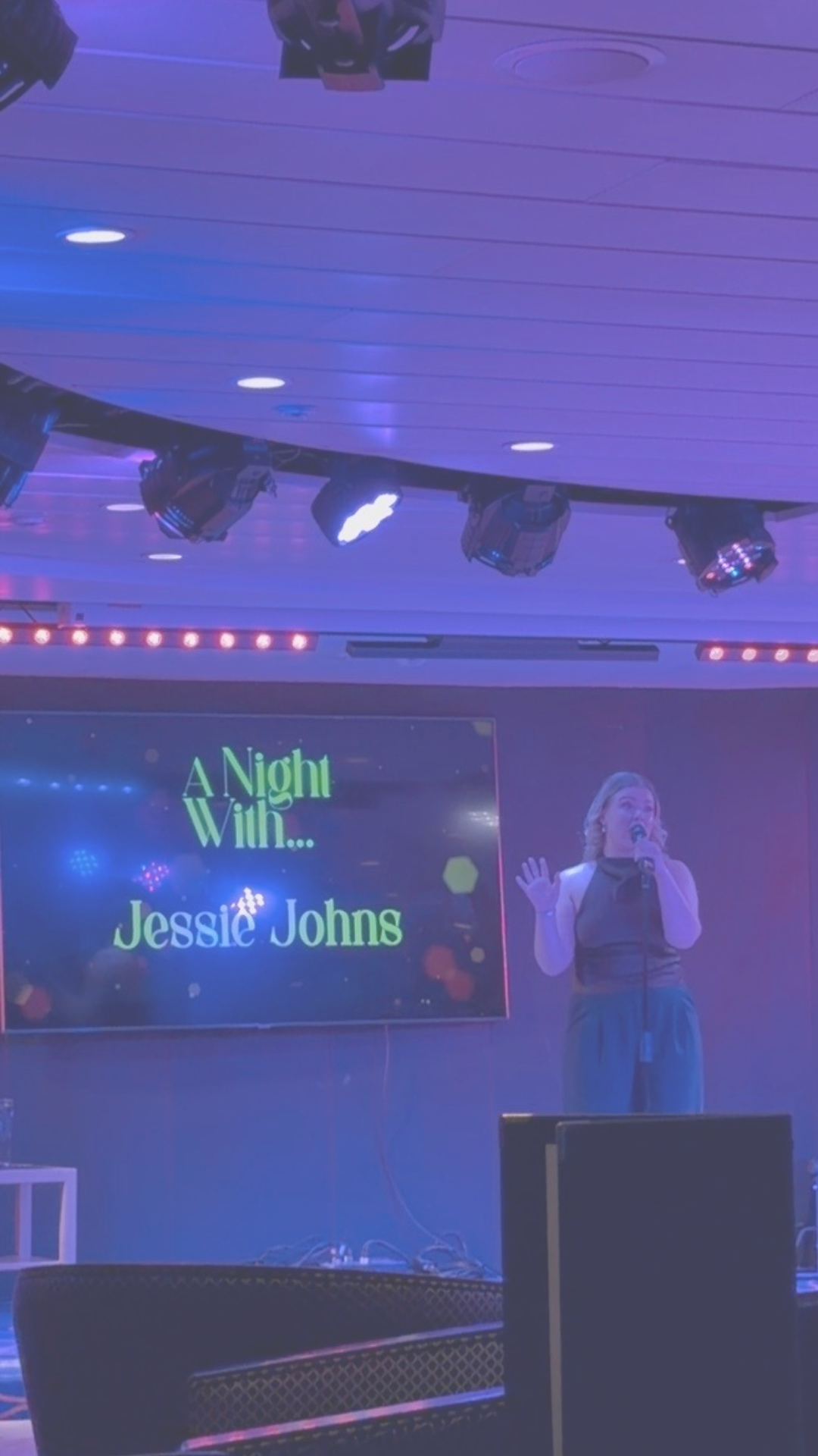 A woman singing on stage with a microphone, in front of a screen that reads "A Night with... Jessie Johns." Stage lights and equipment are visible overhead.