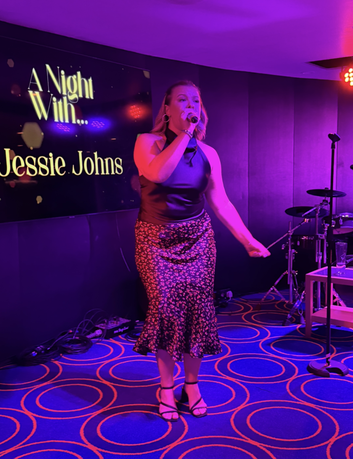 A woman singing into a microphone on stage with purple lighting, in front of a screen that says "A Night with... Jessie Johns." She is wearing a satin top, a floral skirt, and heels.