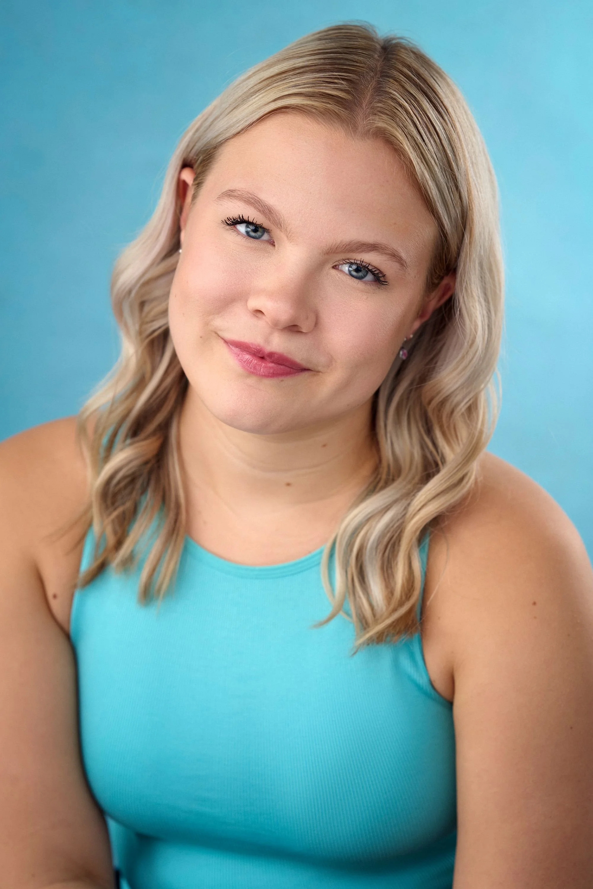 Close-up of a smiling woman with blonde wavy hair, blue eyes, wearing a turquoise sleeveless top, against a blue background.