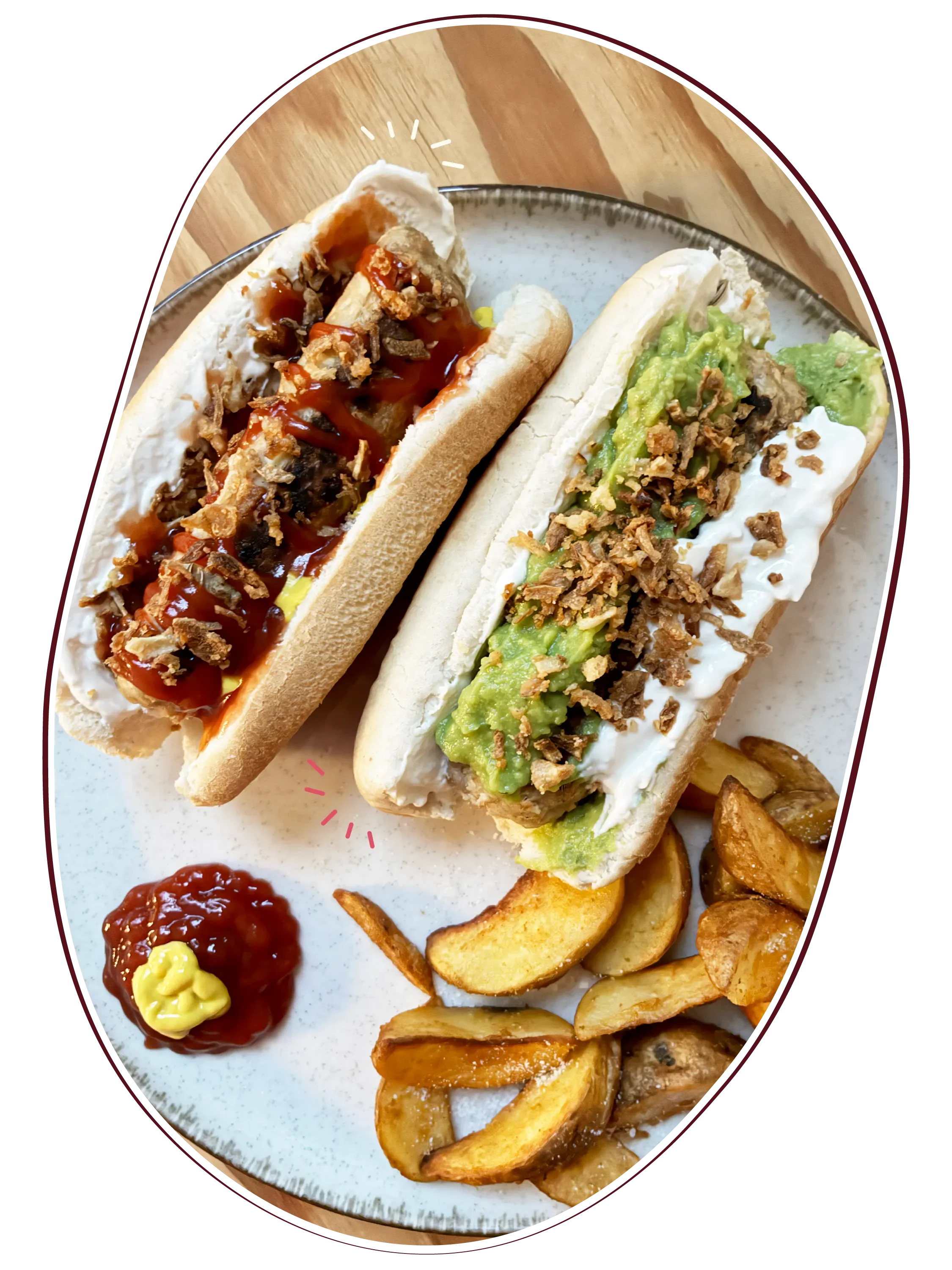 Dos hot dogs veganos con diferentes ingredientes, uno con salchicha, ketchup, mostaza y cebolla frita, y el otro con aguacate, crema y cebolla frita, acompañados de patatas gajo y ketchup con mostaza.