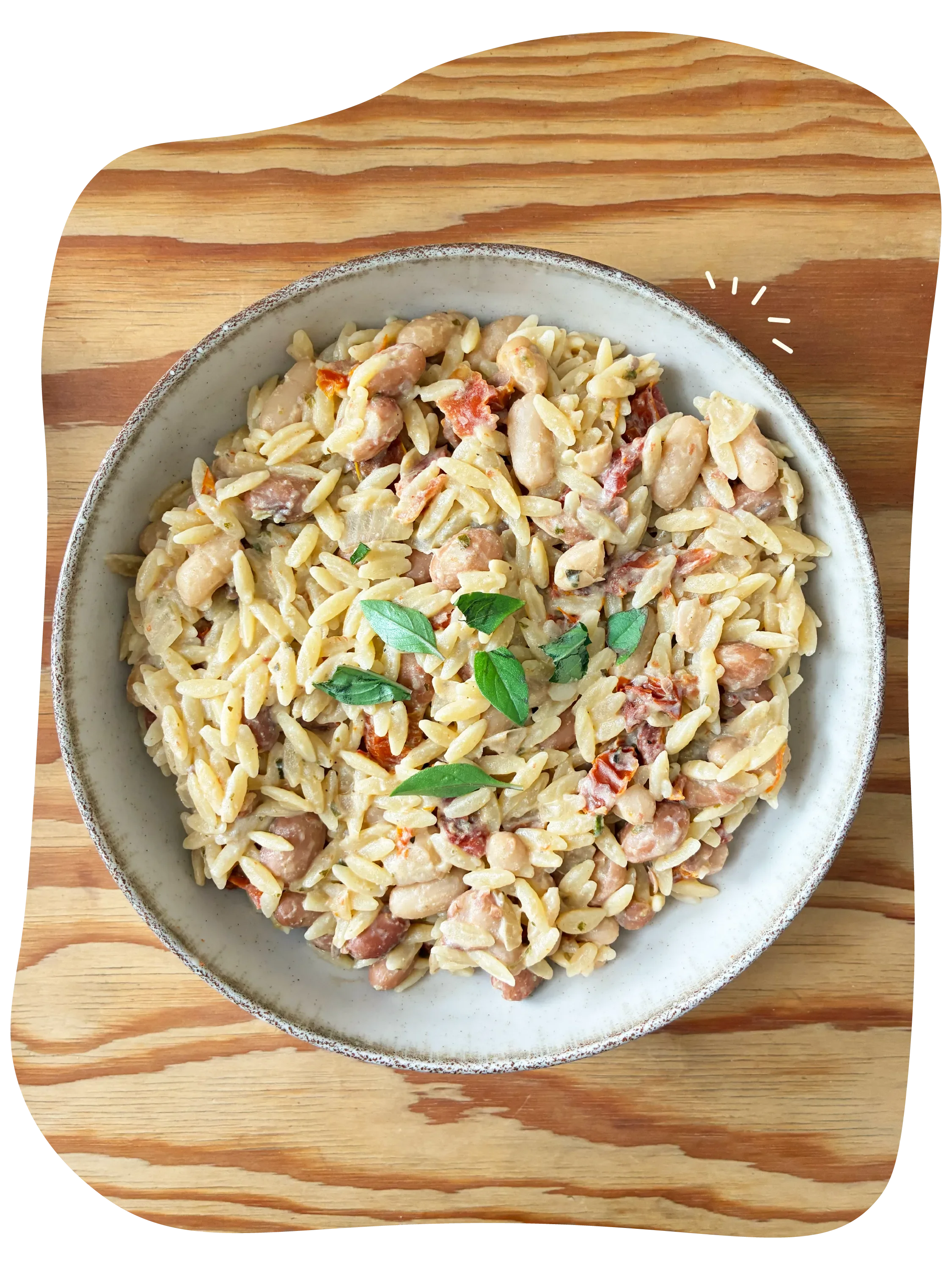 Porción de pasta estilo orzo con alubias pintas, tomates secos, salsa cremosa y hojas de albahaca en un plato de cerámica sobre una tabla de madera.