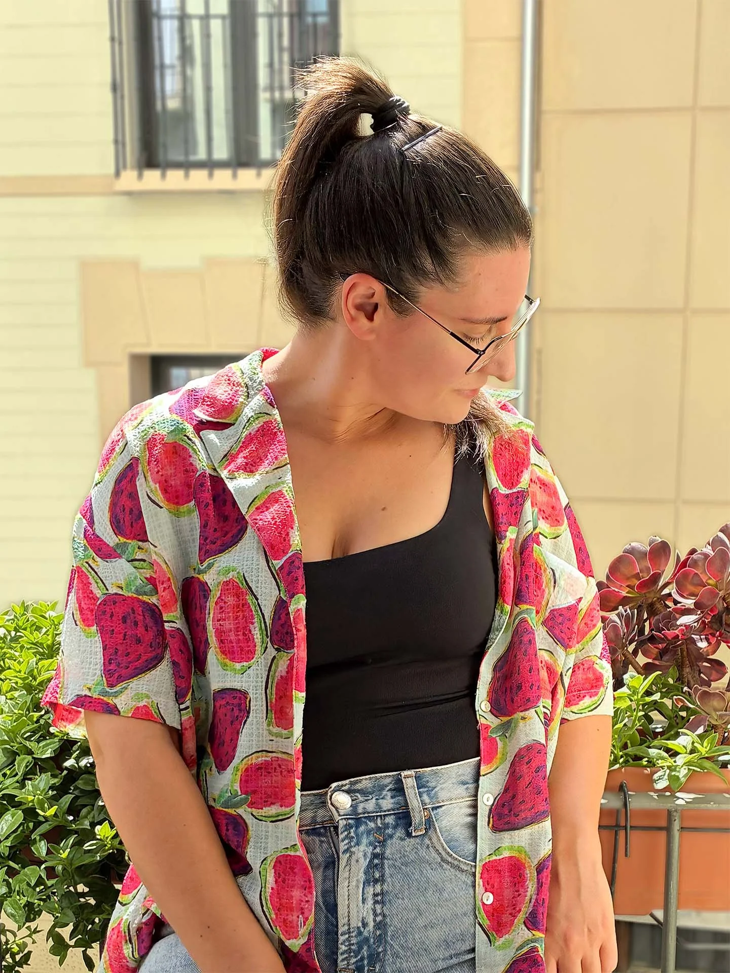 Mujer con gafas, camiseta negra y pantalones vaqueros, lleva una blusa con estampado de sandías en tonos rosas y verdes, y tiene el cabello recogido en una coleta con una pinza, en un ambiente exterior con plantas y fondo de edificio.