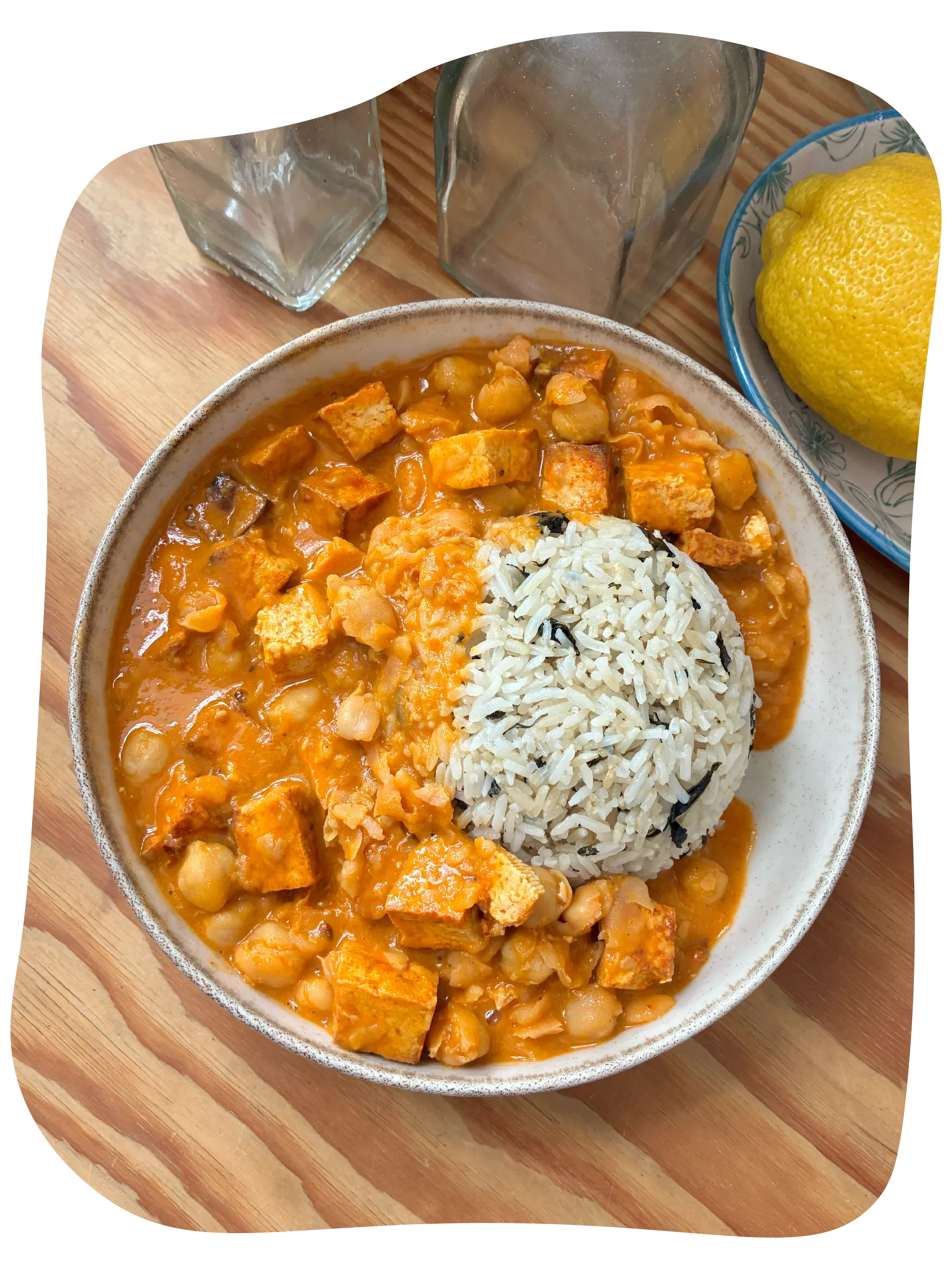 Plato de curry con garbanzos y tofu con arroz blanco y una rodaja de limón en un plato azul, sobre una tabla de madera.