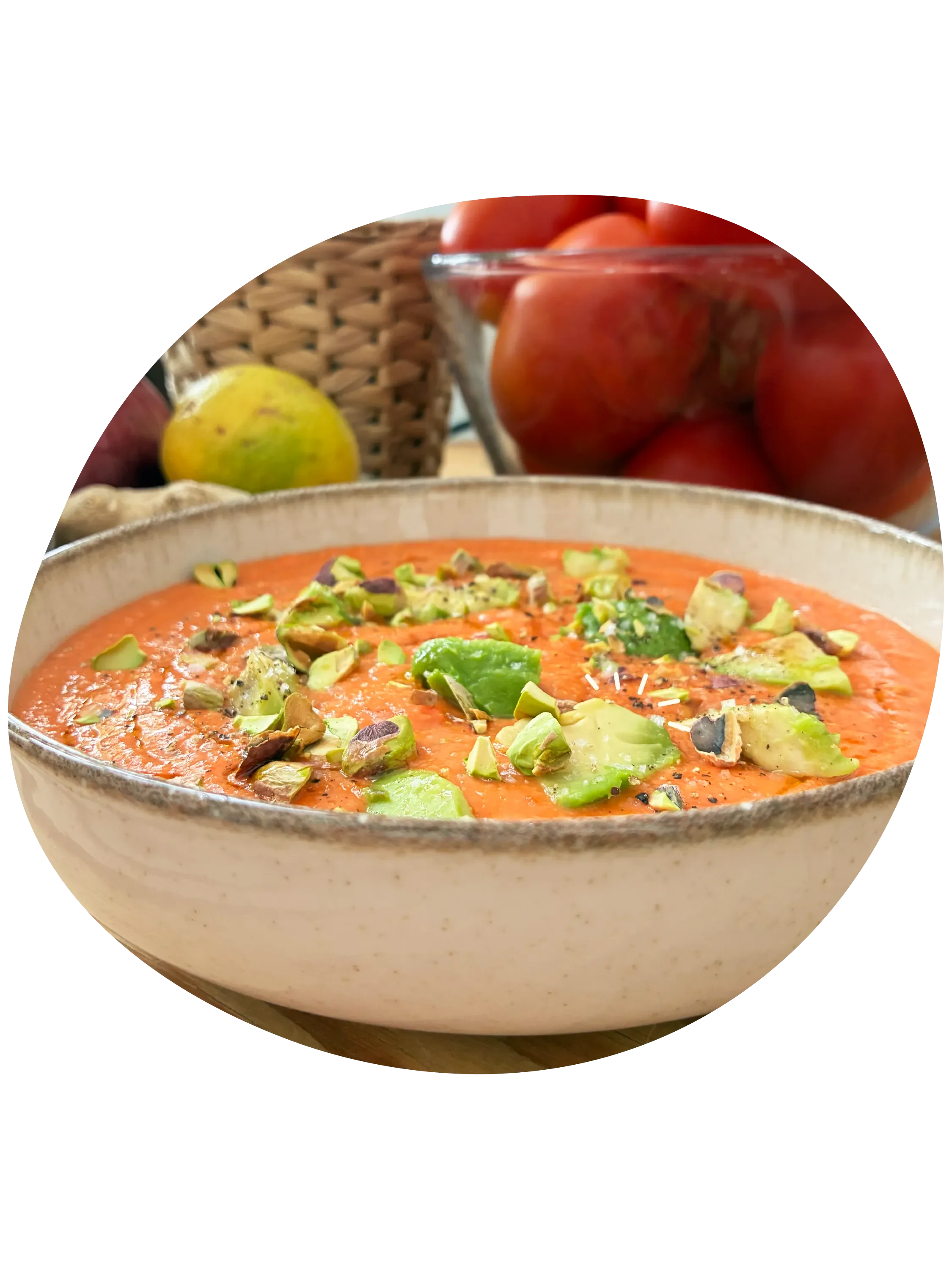 Salmorejo de tomate adornado con pistachos y aguacate en plato de cerámica, con un bol de cristal lleno de tomates y una fruta en una cesta de mimbre en el fondo.