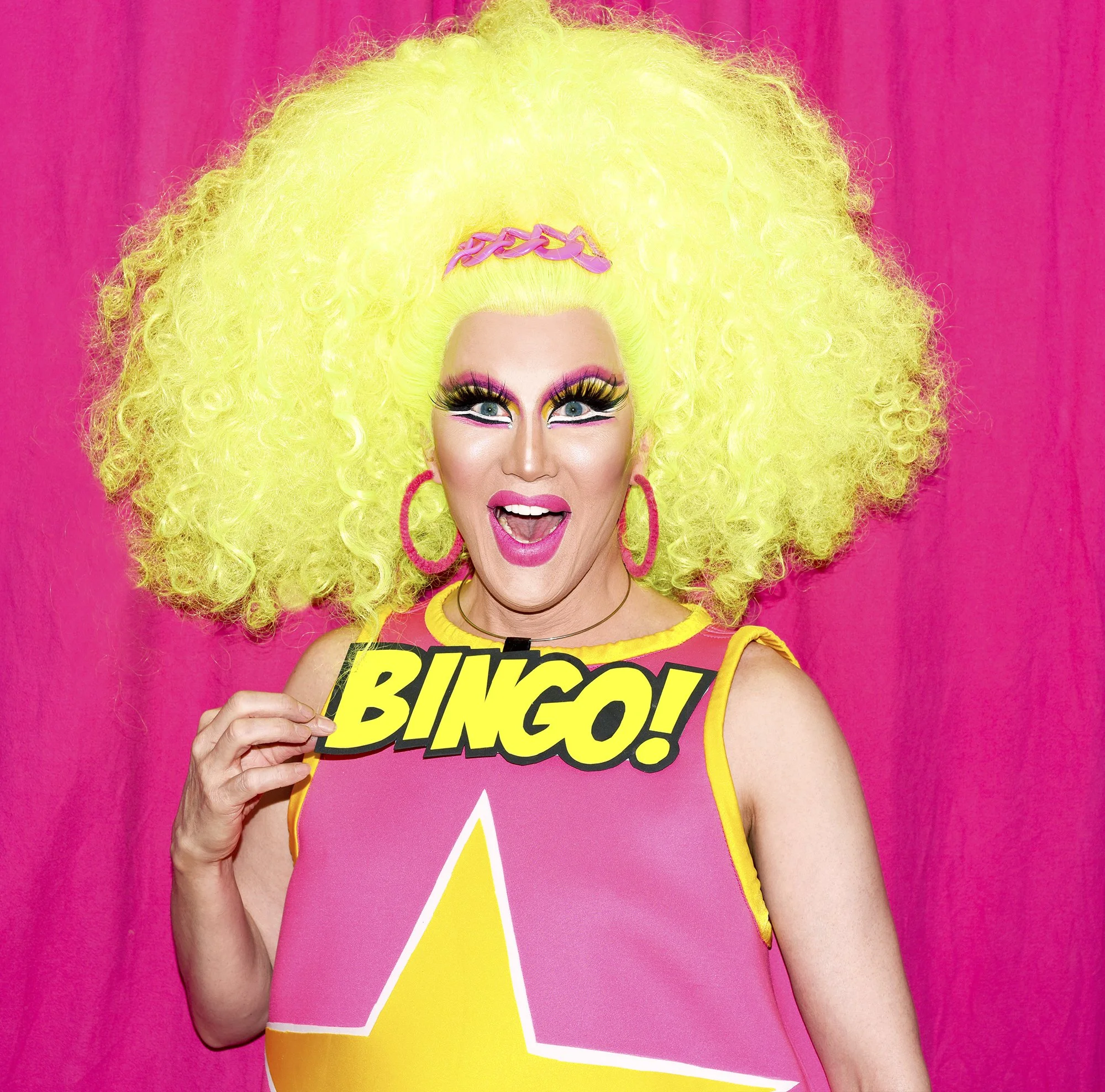 Person with bright yellow curly hair, makeup, and vibrant clothing holding a sign that says 'BINGO!' against a pink background.
