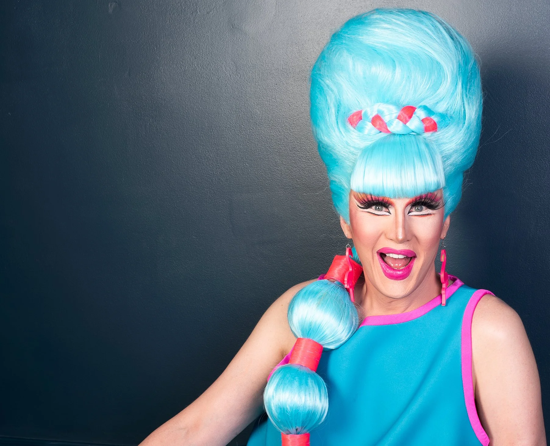 A drag queen with bright blue hair styled in an elaborate beehive, with braids wrapped in red ribbons, smiling and wearing colorful makeup, earrings, and a vibrant blue and pink outfit, standing against a dark gray wall.
