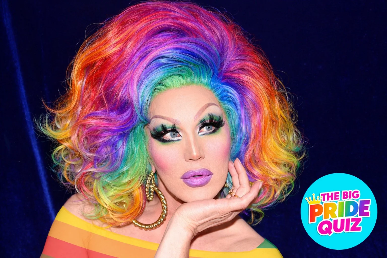 A person with rainbow-colored hair styled in big, voluminous curls, vibrant makeup with green eyeshadow, false eyelashes, pink lipstick, wearing large gold hoop earrings and a rainbow-striped top. There is a blue circle with the text 'The Big Pride Quiz' in rainbow colors in the bottom right corner.