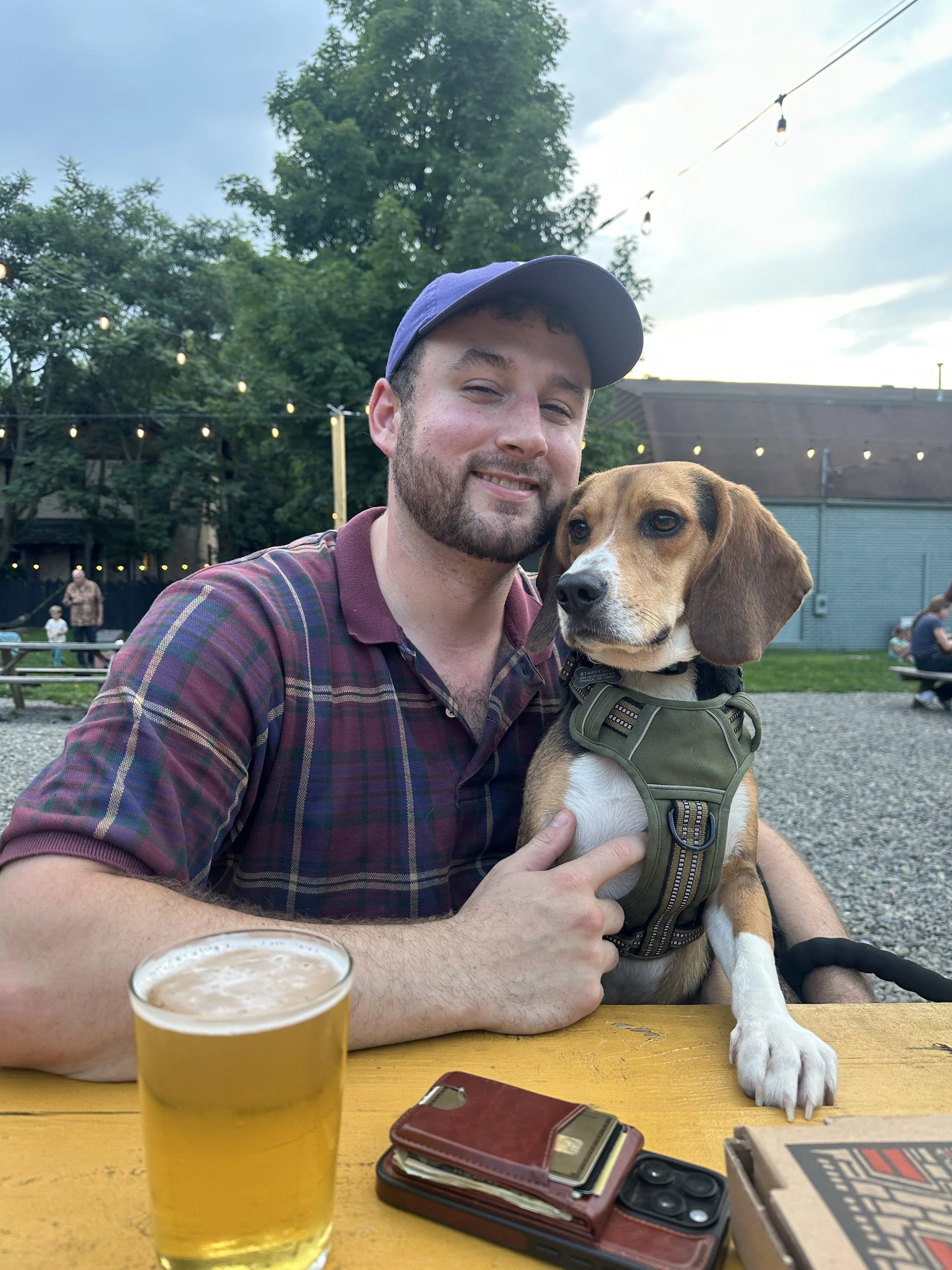 A man with a beard and wearing a blue baseball cap, smiling, sitting at a yellow table with a beer and a dog. The dog is a brown and white beagle wearing a harness, resting its paw on the table, while the man holds it. In the background, there are string lights, trees, and other people at an outdoor gathering.