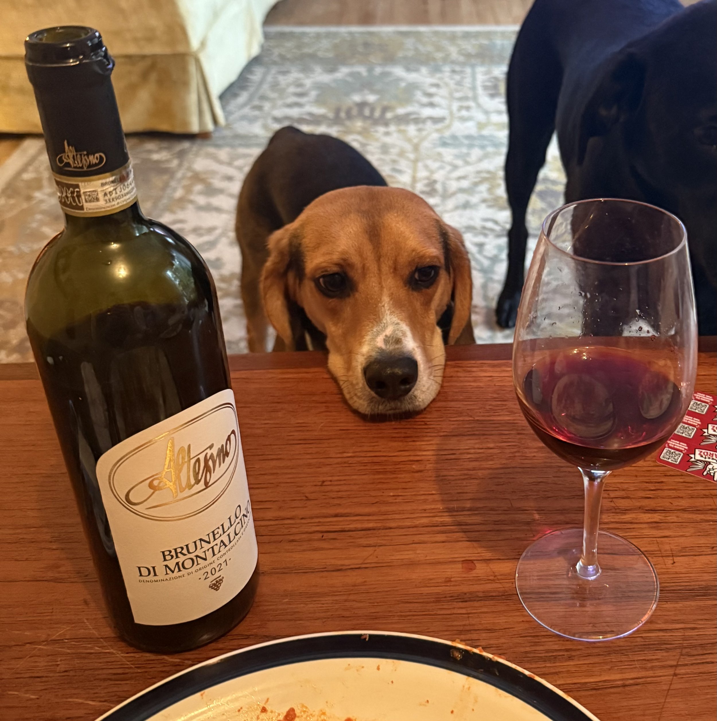 A partially eaten plate of food, a bottle of red wine with a white label, a glass of red wine, and two dogs (one brown and one black) are on a wooden table in a cozy room.