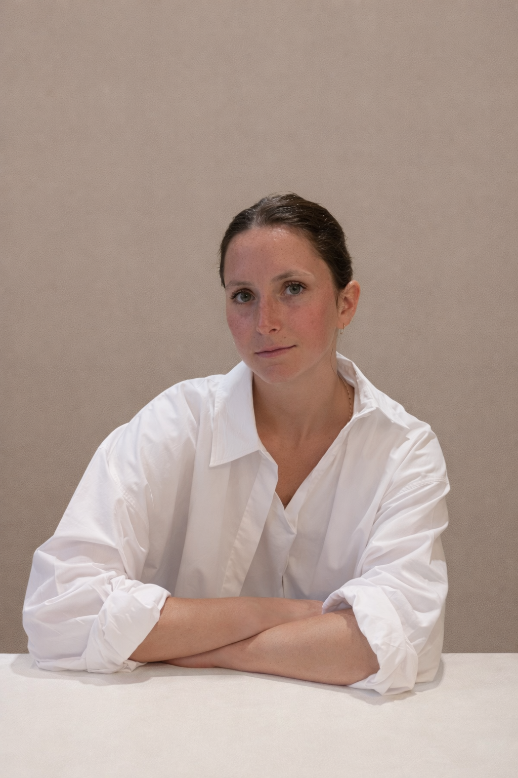 Una donna con capelli castani raccolti, vestita con una camicia bianca, seduta con le braccia incrociate su un tavolo, sfondo beige.