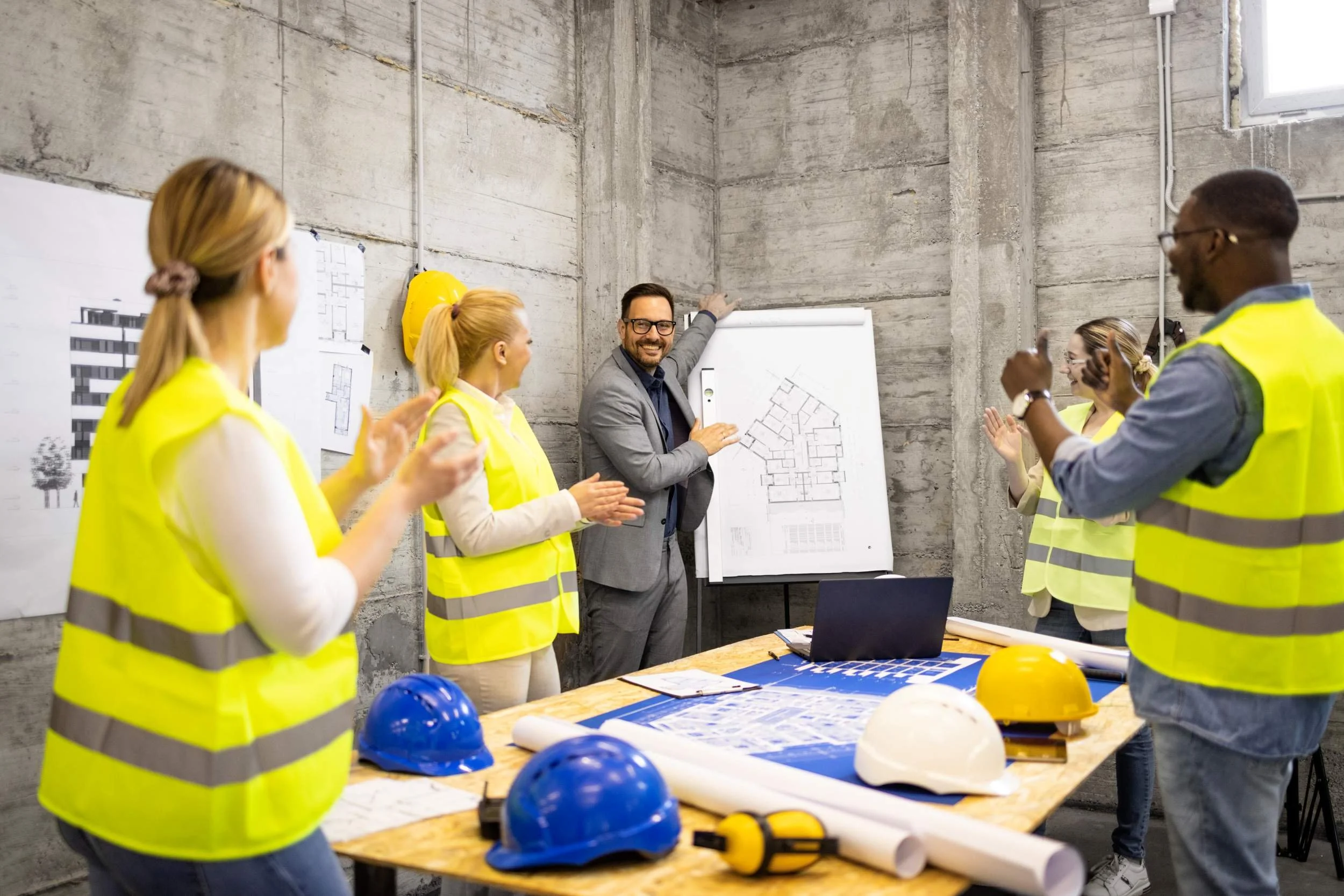 A group of construction professionals, including both men and women, wearing yellow high-visibility vests, are gathered in an unfinished concrete room. They are smiling and clapping while a man in glasses and a gray suit presents blueprints. Construction helmets, blueprints, and other planning materials are on the table.