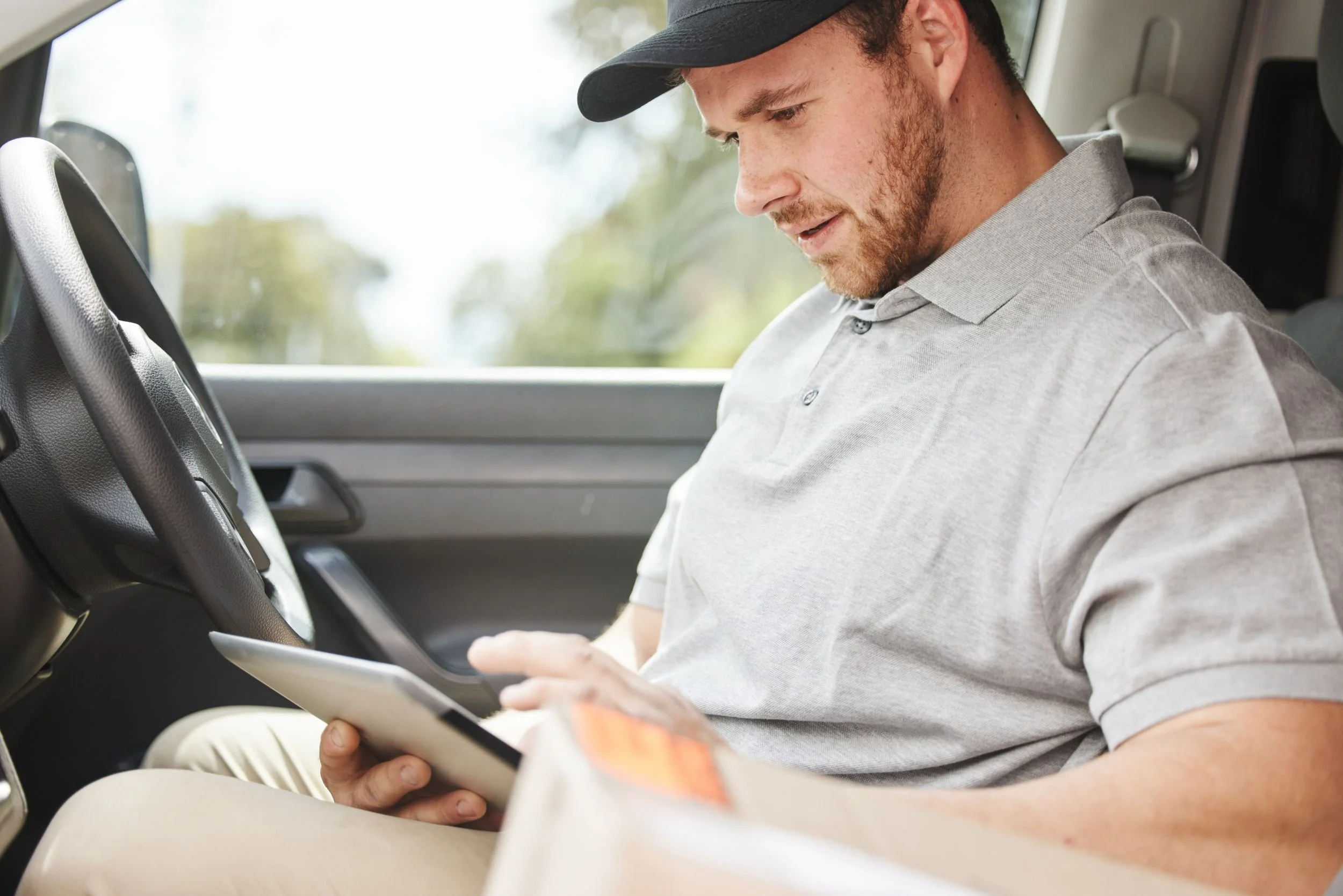 A young man with a beard and mustache wearing a gray polo shirt and black cap sitting in a vehicle, looking at his smartphone.