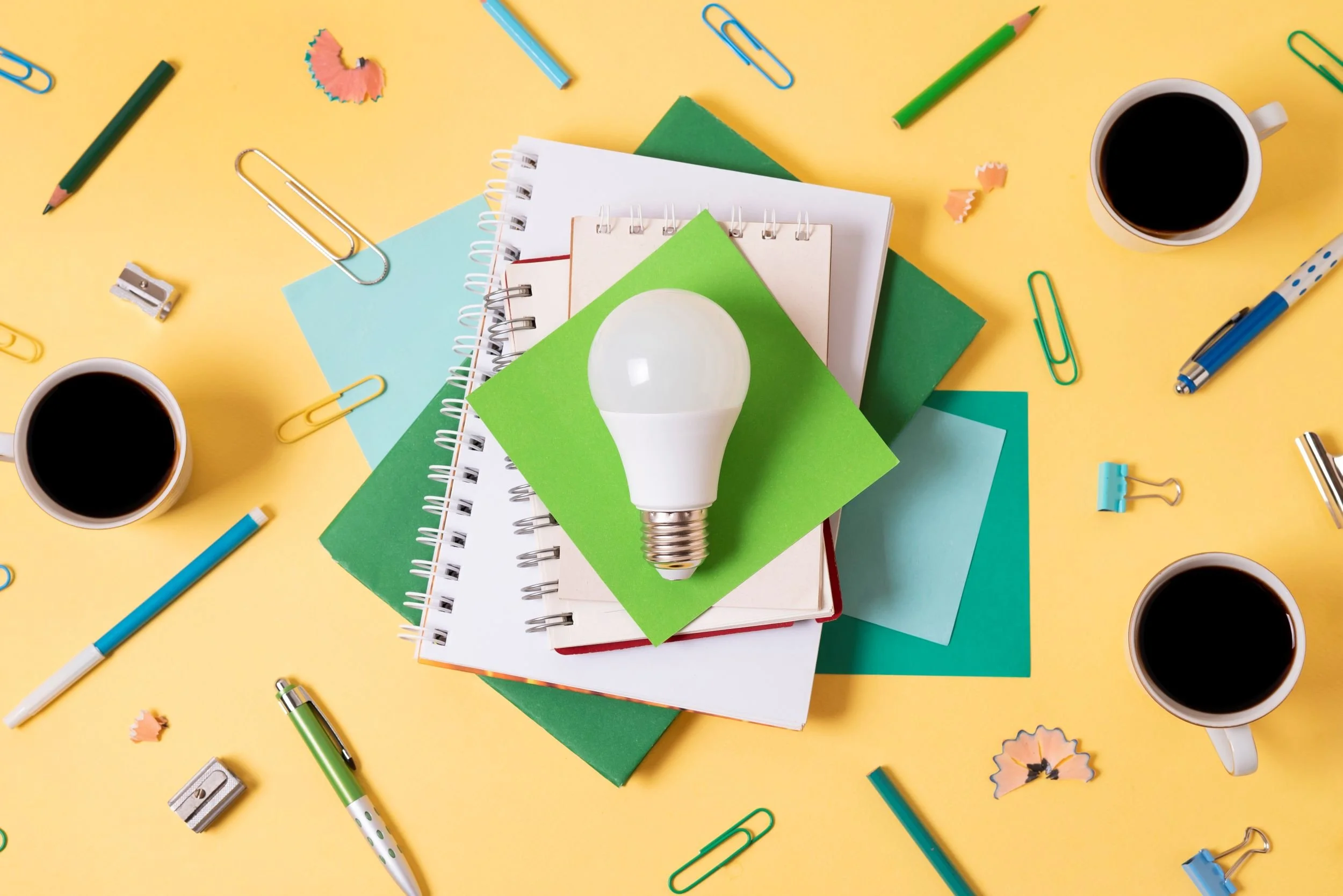 An overhead view of a desk with stationery and notebooks on a yellow background. There are cups of coffee, colored pencils, paper clips, a sharpener, pencil shavings, and a light bulb on top of the notebooks.