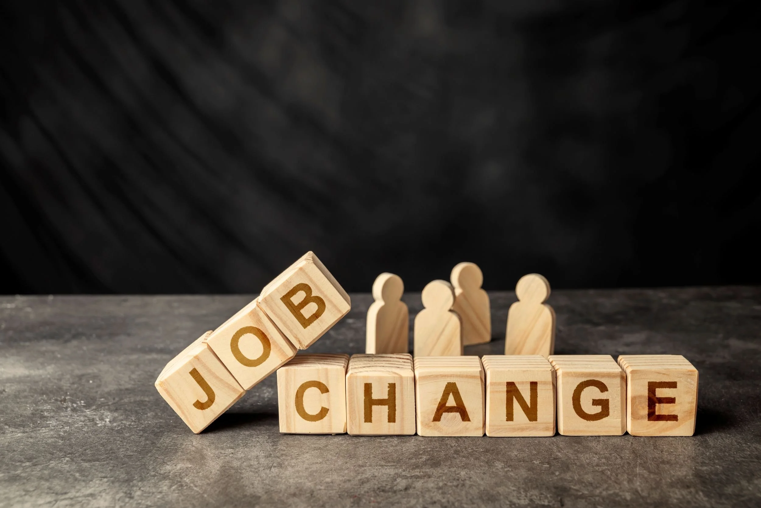 Wooden blocks spelling the word 'CHANGE' on a gray surface, with a tilted block showing parts of the word 'JOB' falling off from the left side, and small wooden human figures in the background against a black backdrop.