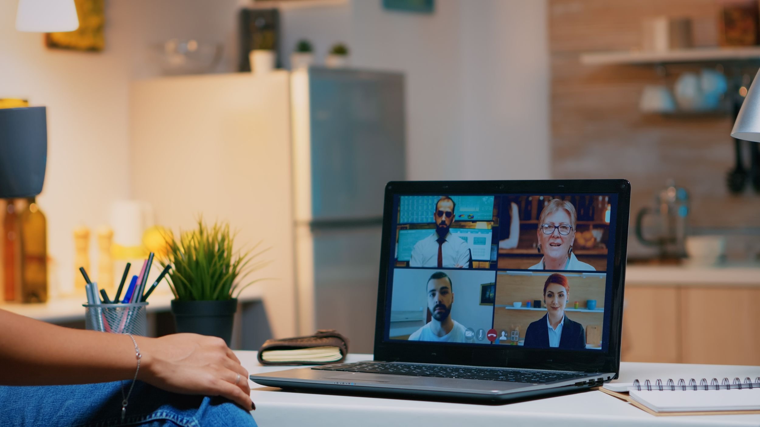 A person participating in a virtual video call with four other people on a laptop screen. The setting is a modern room with a lamp, potted plant, notebooks, and colorful pens on the desk.