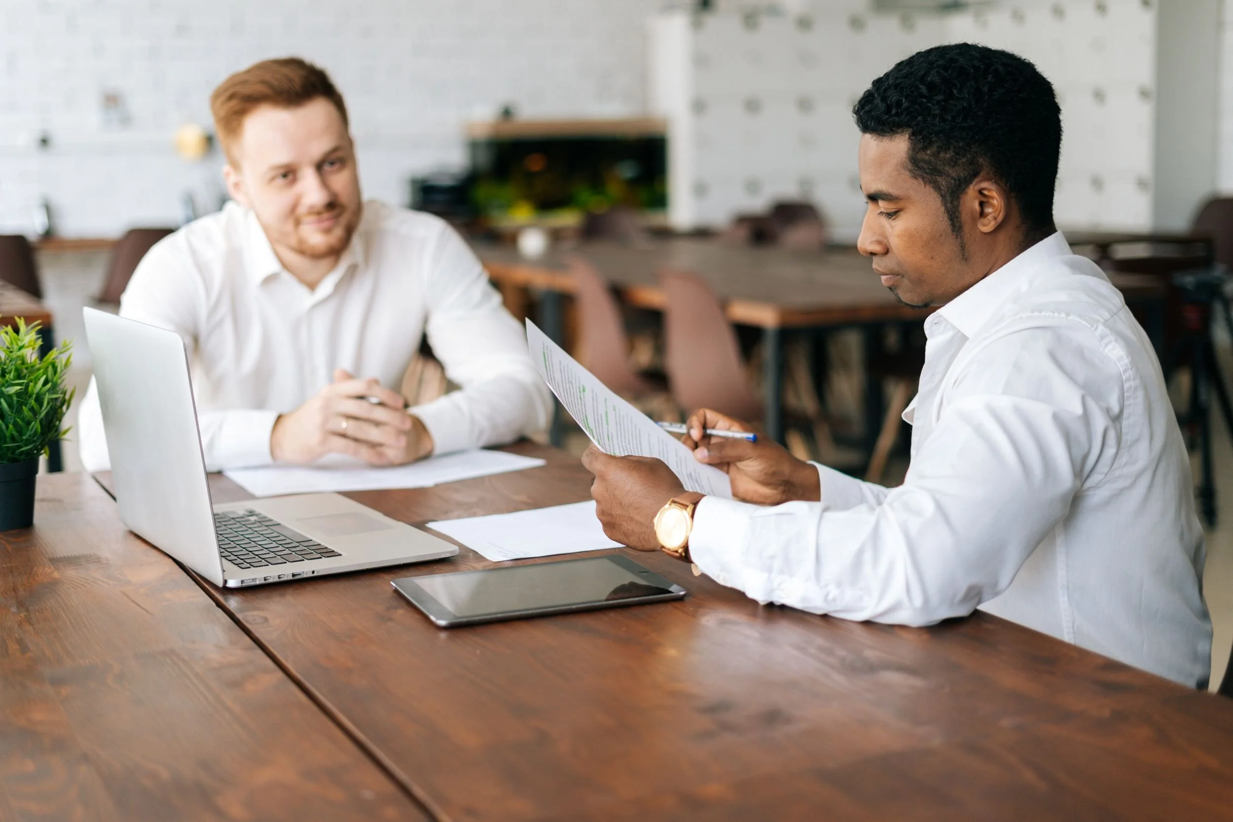 Two men in white shirts sitting at a wooden table in a modern office, discussing documents, with laptops and a potted plant on the table.