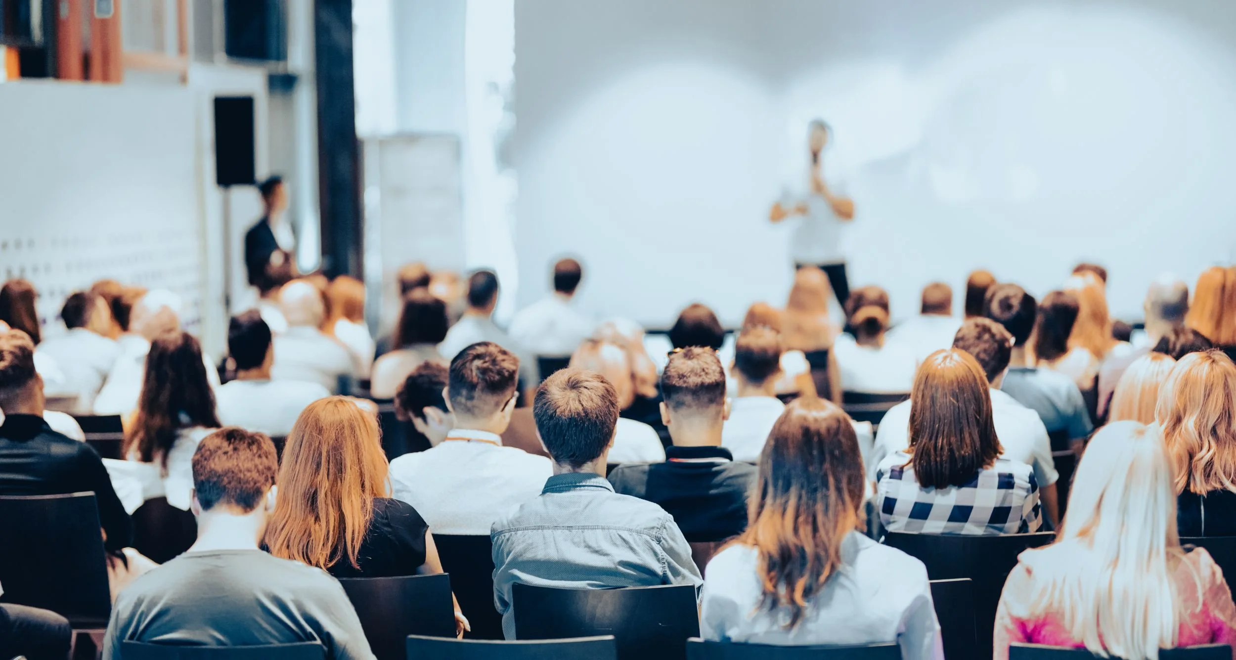 A large group of people seated in chairs facing a whiteboard with a speaker standing in front. The audience, with varied hair colors and styles, appears to be attentively listening to the presentation.