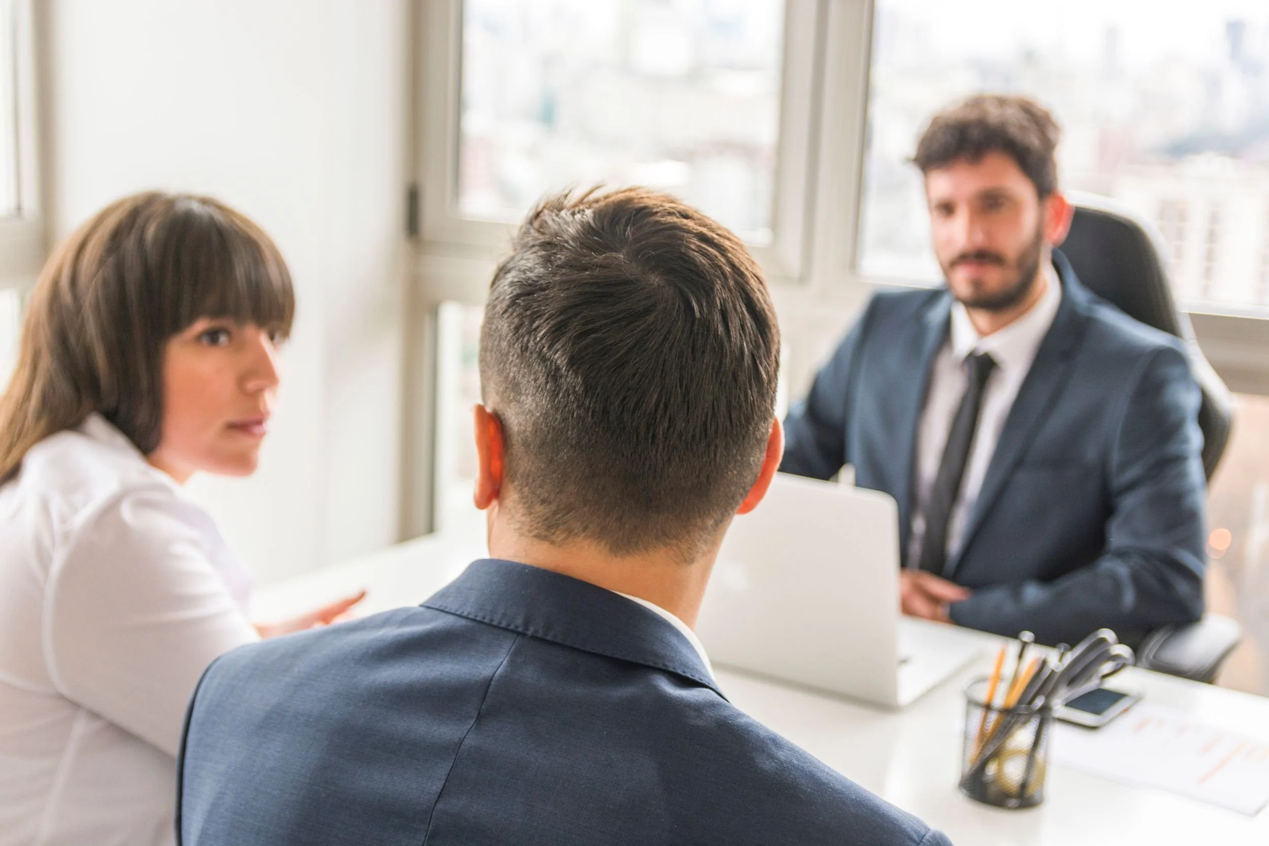 Four people in a professional meeting room engaged in a discussion, with a man in a suit on the right and three others, including a woman with brown hair, facing him.
