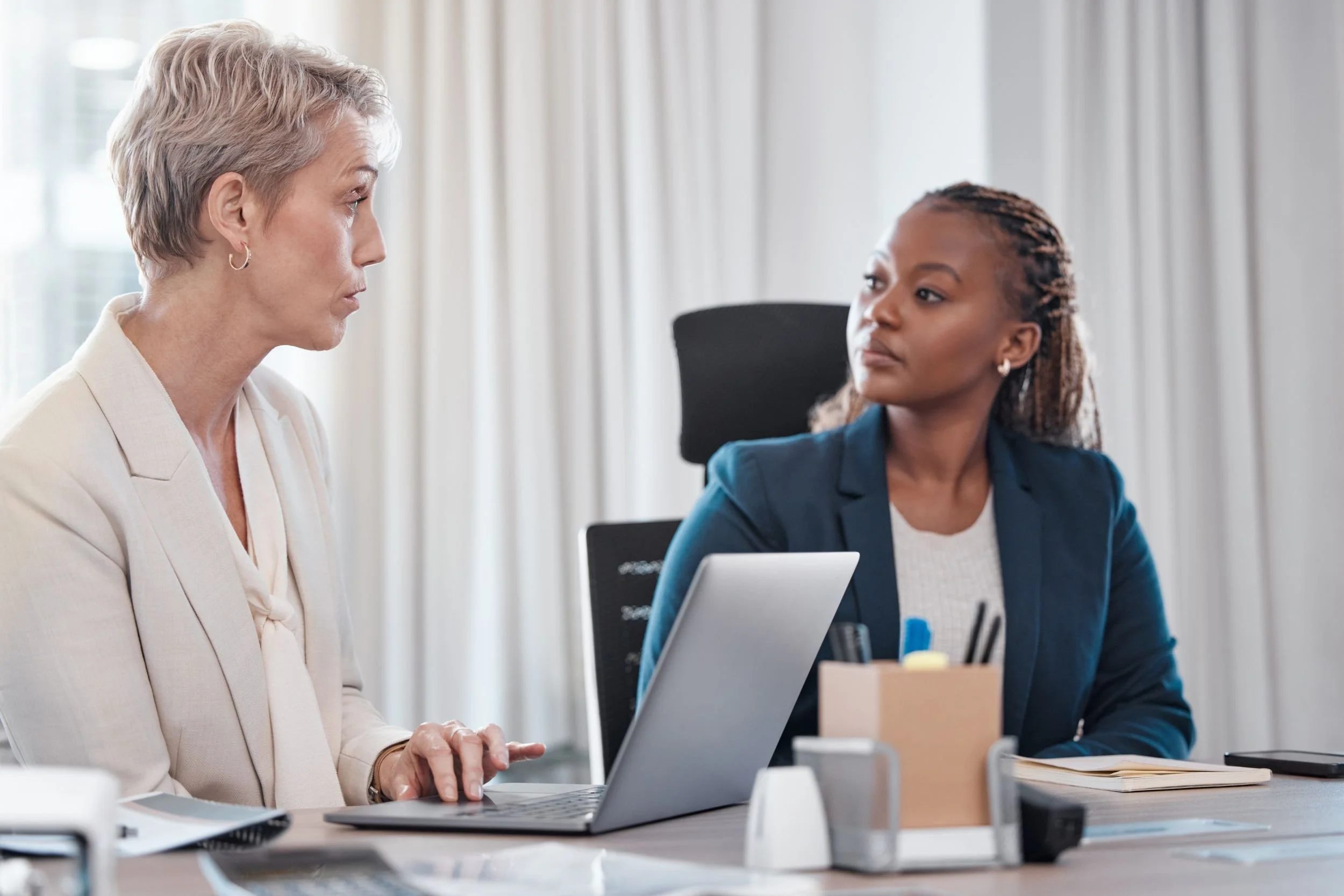 Two women engaged in a serious conversation in a modern office, with one woman working on a laptop and the other listening attentively.