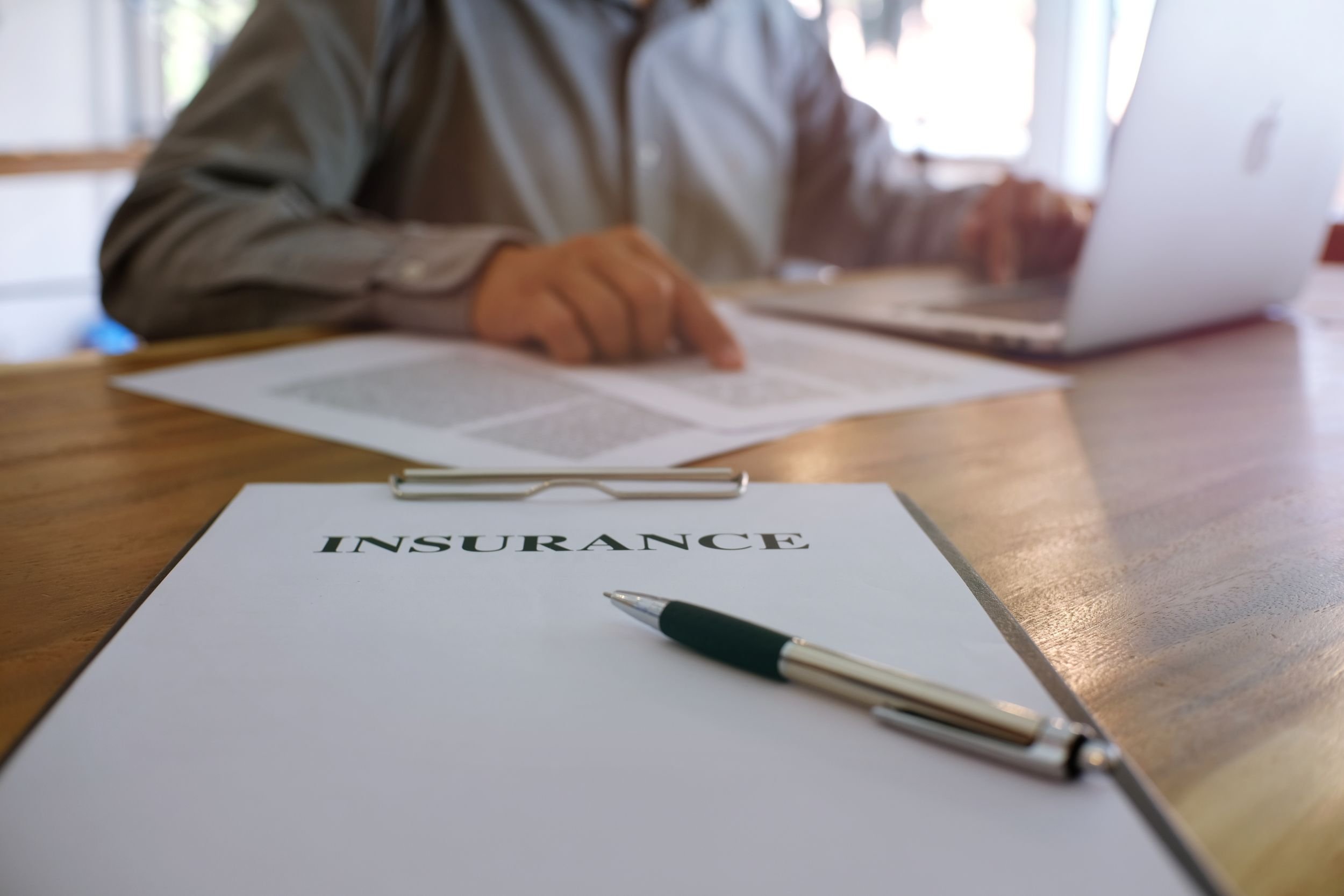 A desk scene with an insurance document and a pen in the foreground, and a person in a shirt working on a laptop in the background, with natural light streaming through a window.