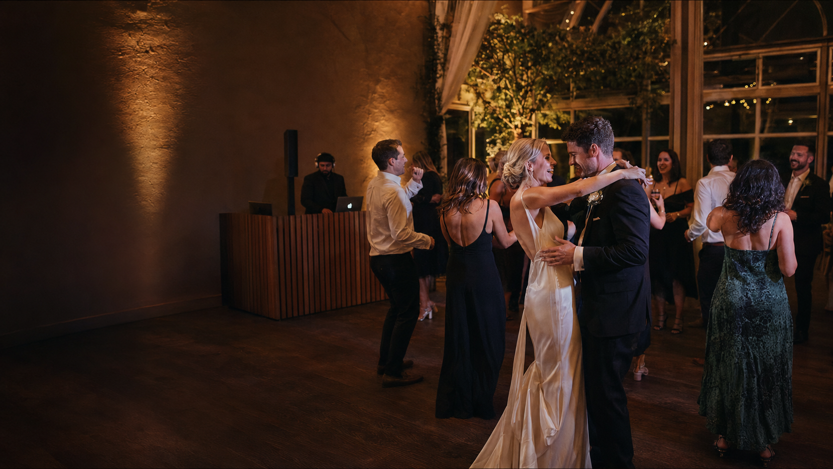 Couple dancing together at a wedding reception, surrounded by guests, in a dimly lit elegant venue with large windows and warm lighting.