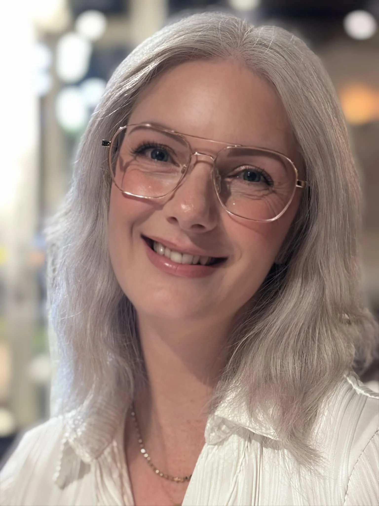 Close-up of a smiling older woman with long gray hair, glasses, and a pearl necklace, wearing a white textured blouse.