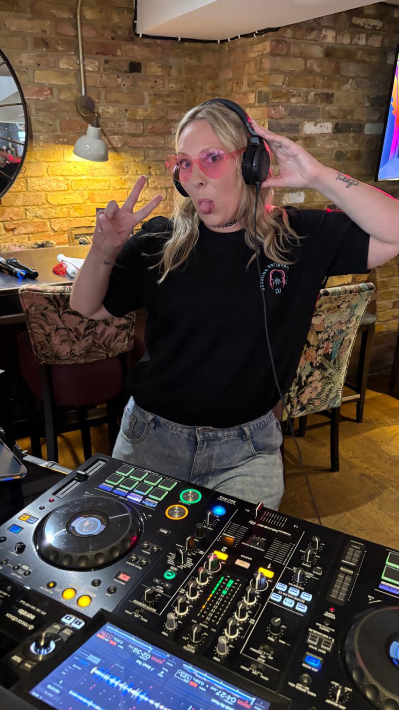 A woman with blonde hair wearing pink sunglasses and headphones making a peace sign and sticking out her tongue, standing behind DJ equipment in a room with a brick wall background.