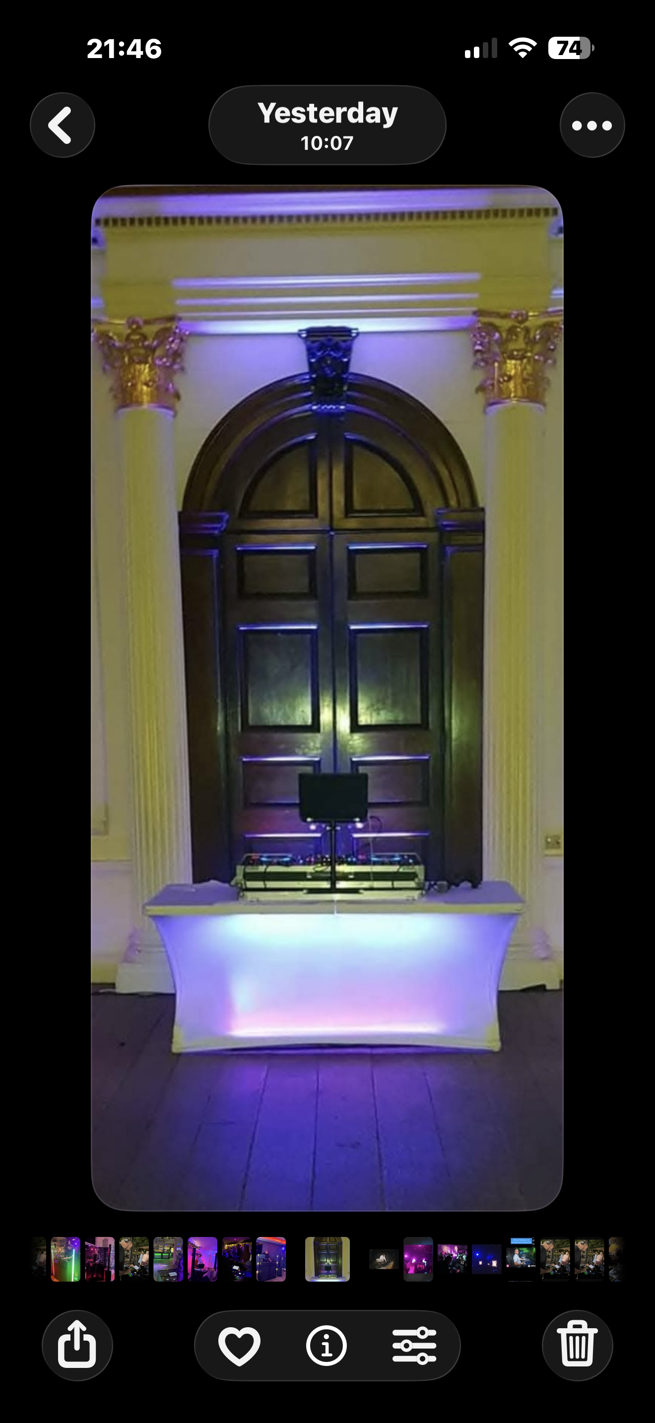A DJ setup with a laptop and DJ equipment on a table illuminated with colorful LED lights, inside a room with ornate columns and a large wooden arched door in the background.