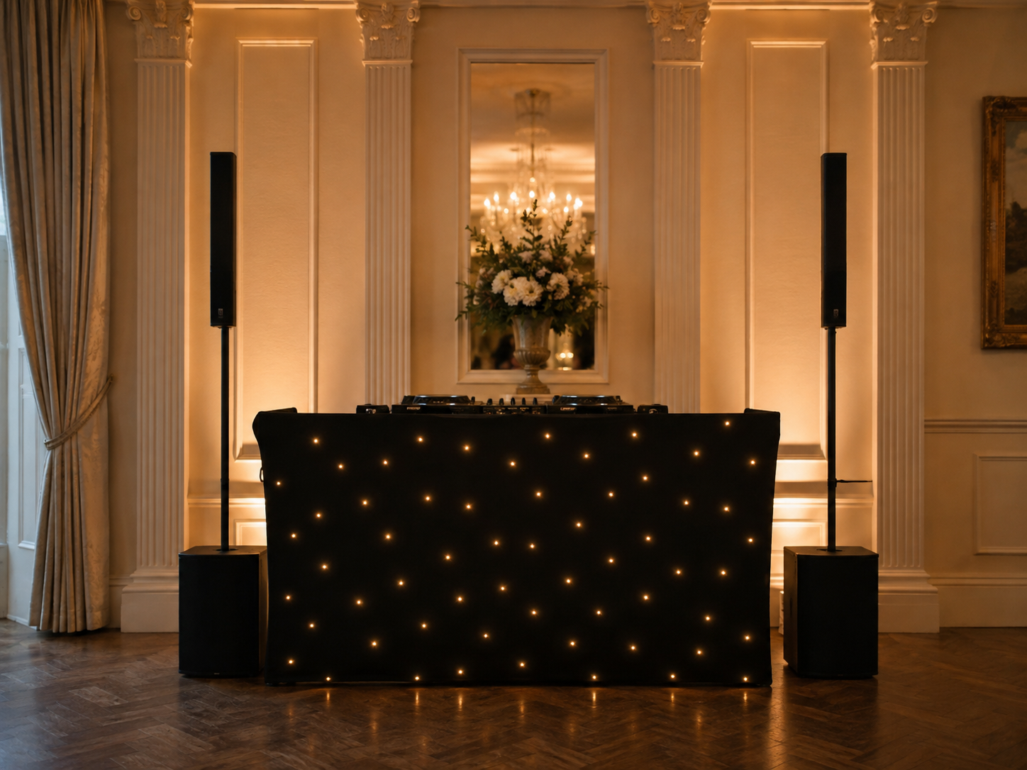 A DJ setup with a black illuminated desk, speakers, and a floral arrangement in a large vase reflected in a mirror behind, all in an elegant room with ornate curtains and chandeliers.