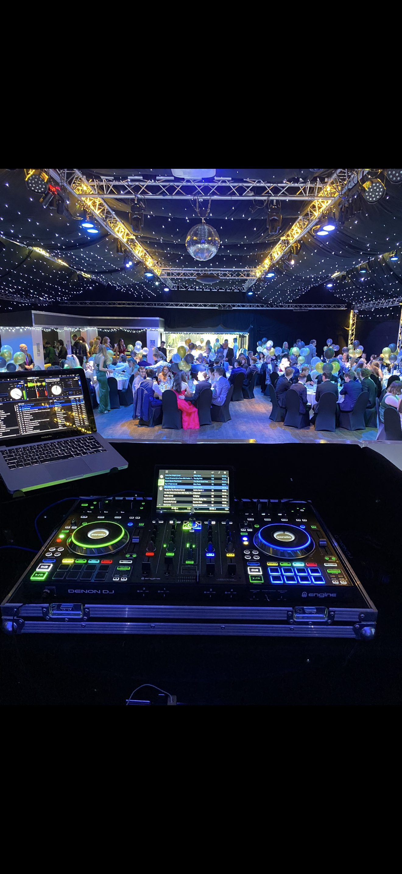 A DJ's equipment setup overlooking a large banquet hall filled with guests seated at decorated tables with balloons, under ceiling draped with string lights and a disco ball.