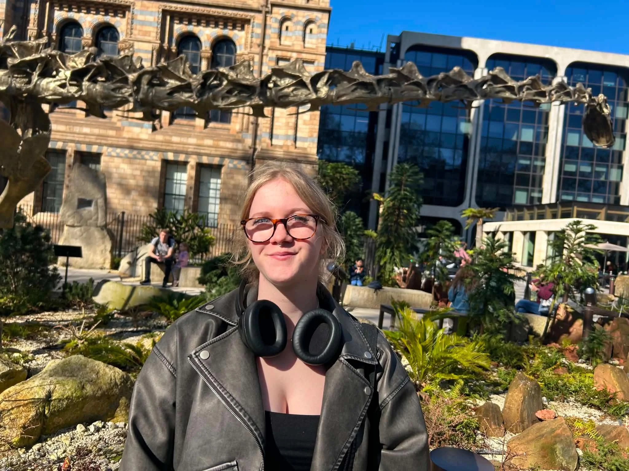 A woman with glasses and a leather jacket taking a selfie outdoors in front of a decorative sculpture of a dragon made of metal with buildings and people in the background.